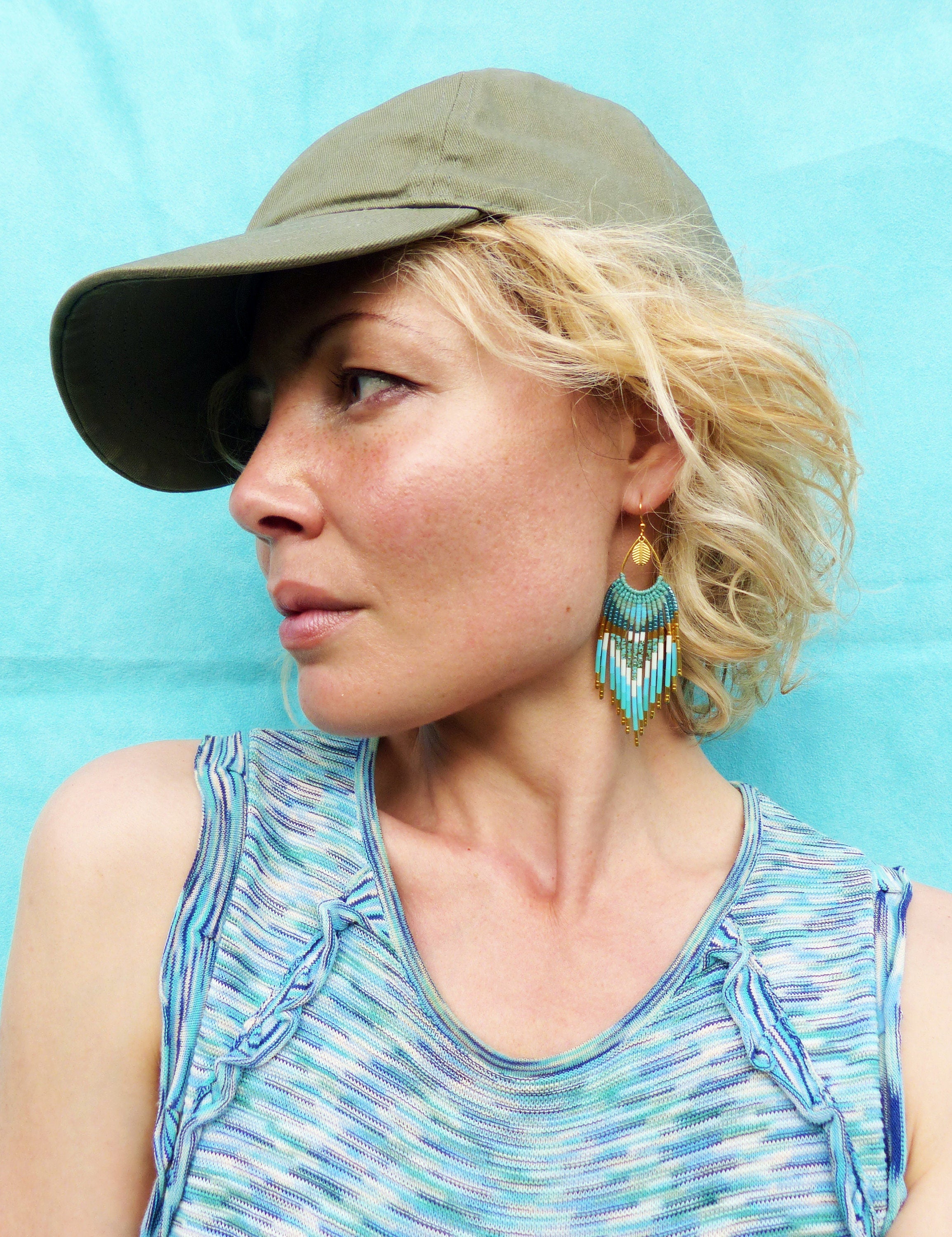 Peut inclure: Une femme porte une casquette de baseball verte et un haut à motifs bleus et blancs. Elle porte des boucles d'oreilles en perles turquoise et dorées. L'arrière-plan est bleu clair uni.