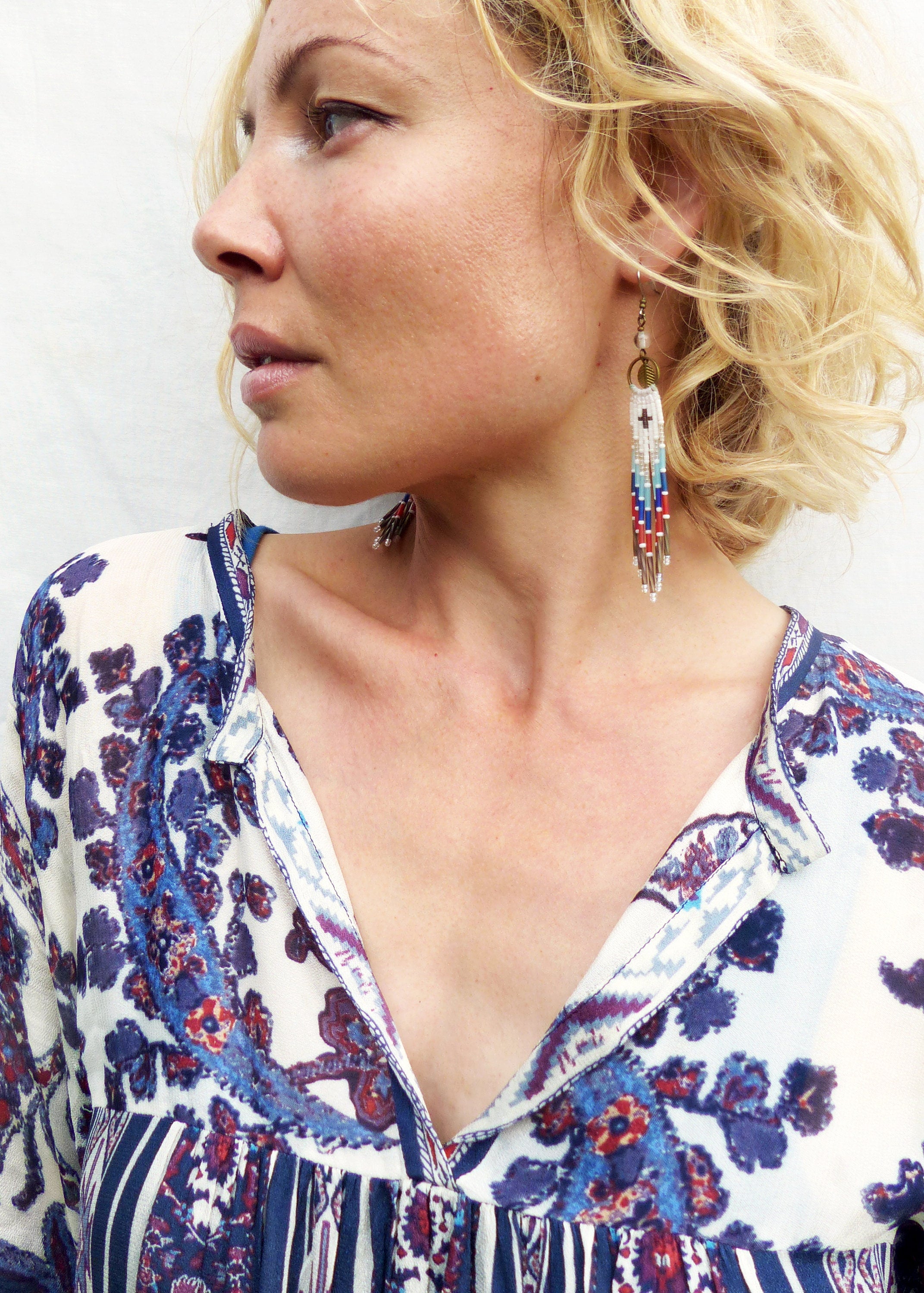 Peut inclure: Une femme aux cheveux blonds portant un chemisier à motif blanc et bleu avec un col en V. Elle porte de longues boucles d'oreilles en perles avec des perles blanches, bleues, rouges et dorées.