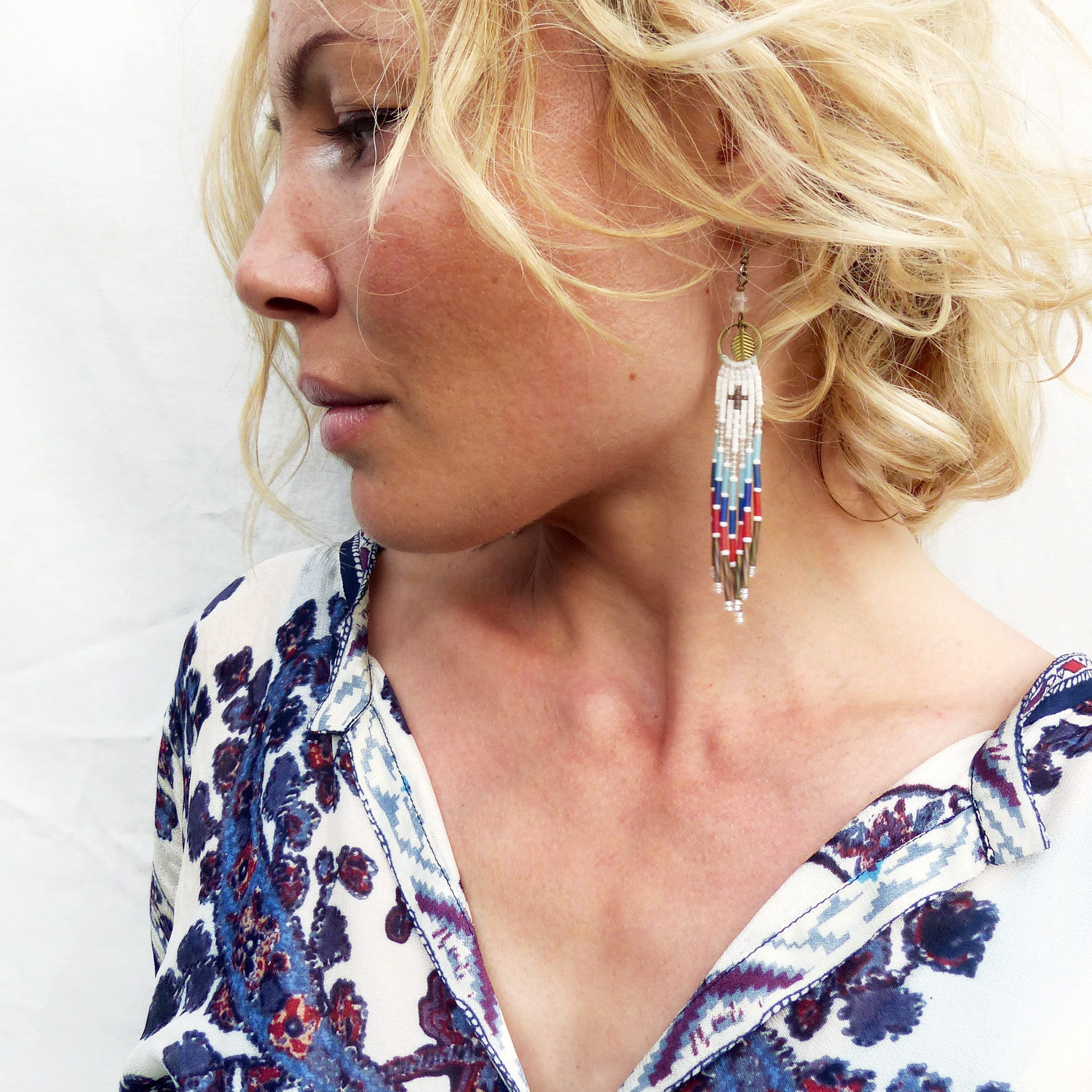 Peut inclure: Une femme aux cheveux blonds portant un chemisier à motifs blancs et bleus avec de longues boucles d'oreilles en perles. Les boucles d'oreilles sont blanches avec des accents rouges, bleus et dorés.