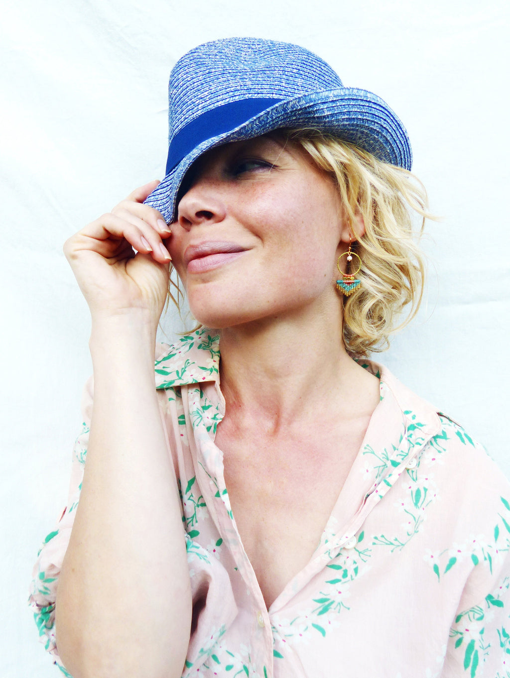 Peut inclure: Un chapeau de paille bleu avec un ruban bleu foncé. Le chapeau est porté de biais. La personne porte une chemise rose à motifs floraux et des boucles d'oreilles dorées avec des perles turquoise et blanches.
