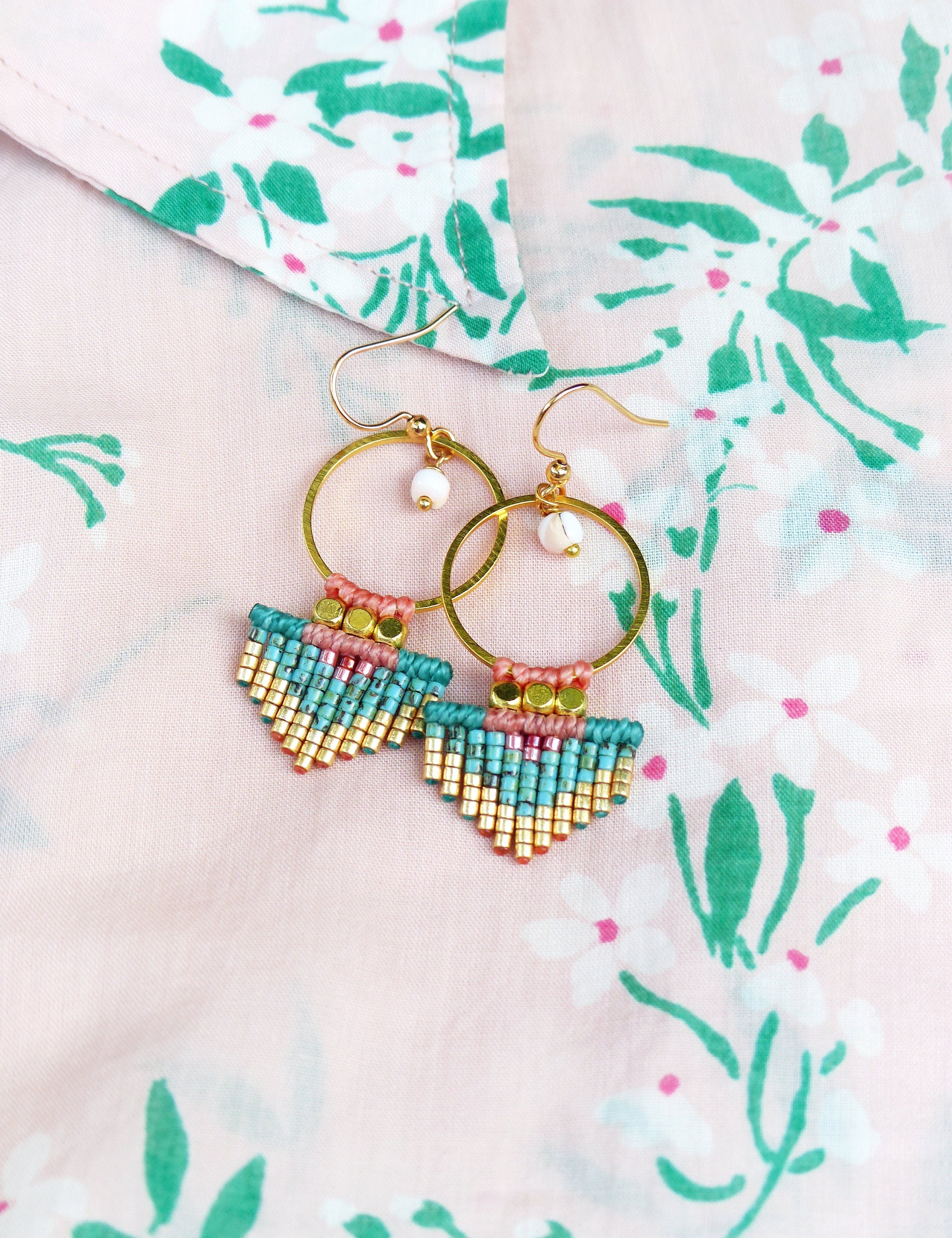 Peut inclure: Une paire de boucles d'oreilles créoles dorées avec un motif géométrique perlé. Les boucles d'oreilles présentent des perles turquoise, roses et dorées, avec une petite perle blanche suspendue à la créole. Les boucles d'oreilles reposent sur un tissu floral rose.