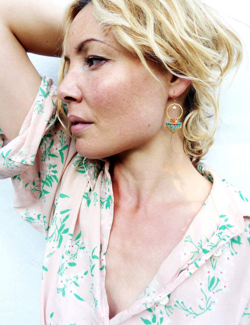 Peut inclure: Une femme portant des boucles d'oreilles dorées avec des détails de perles colorées. Les boucles d'oreilles ont un design circulaire avec une petite perle et une forme de demi-lune avec des perles turquoise, rouges et jaunes. La femme porte une chemise rose à fleurs.