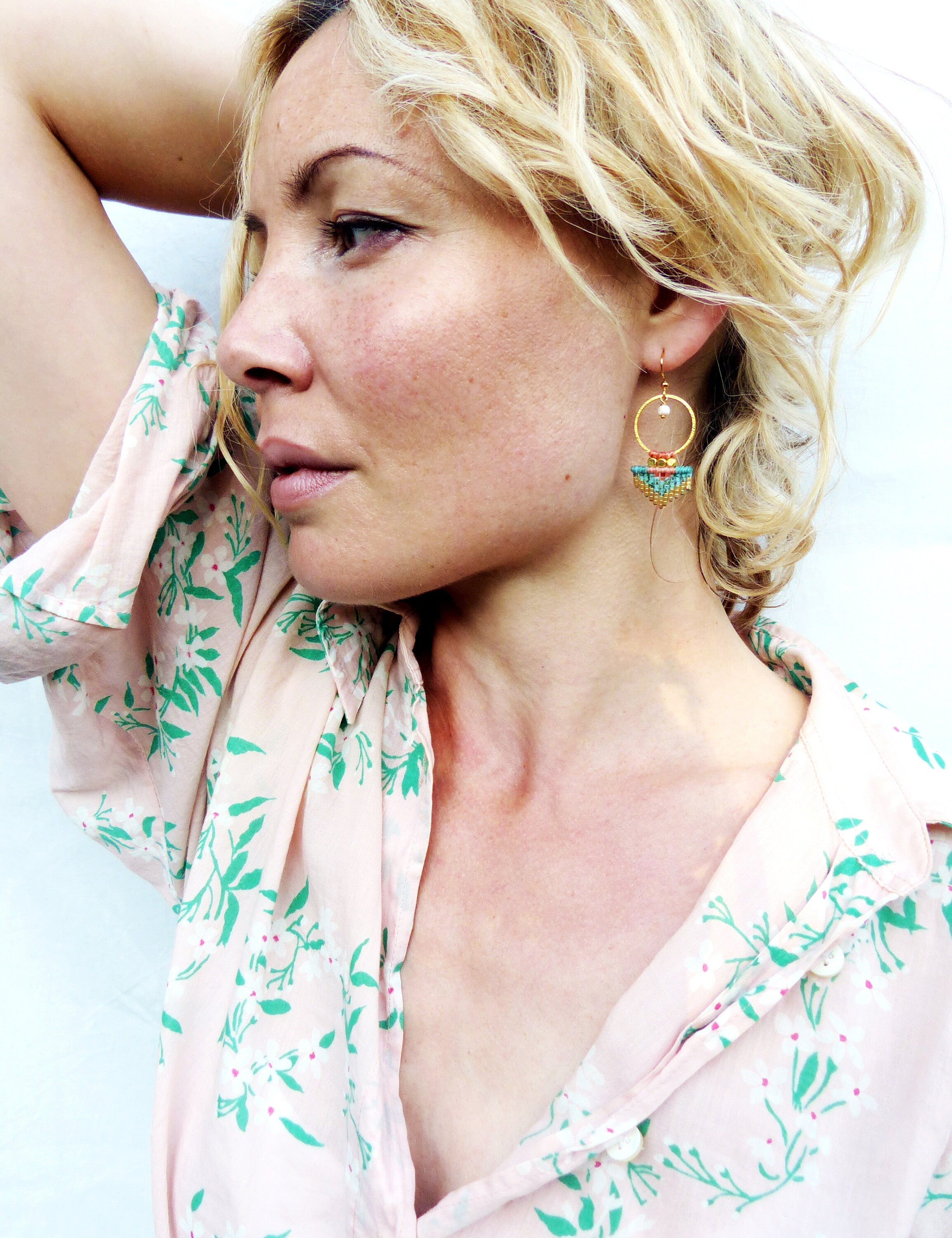 Peut inclure: Une femme portant des boucles d'oreilles dorées avec des détails de perles colorées. Les boucles d'oreilles ont un design circulaire avec une petite perle et une forme de demi-lune avec des perles turquoise, rouges et jaunes. La femme porte une chemise rose à fleurs.