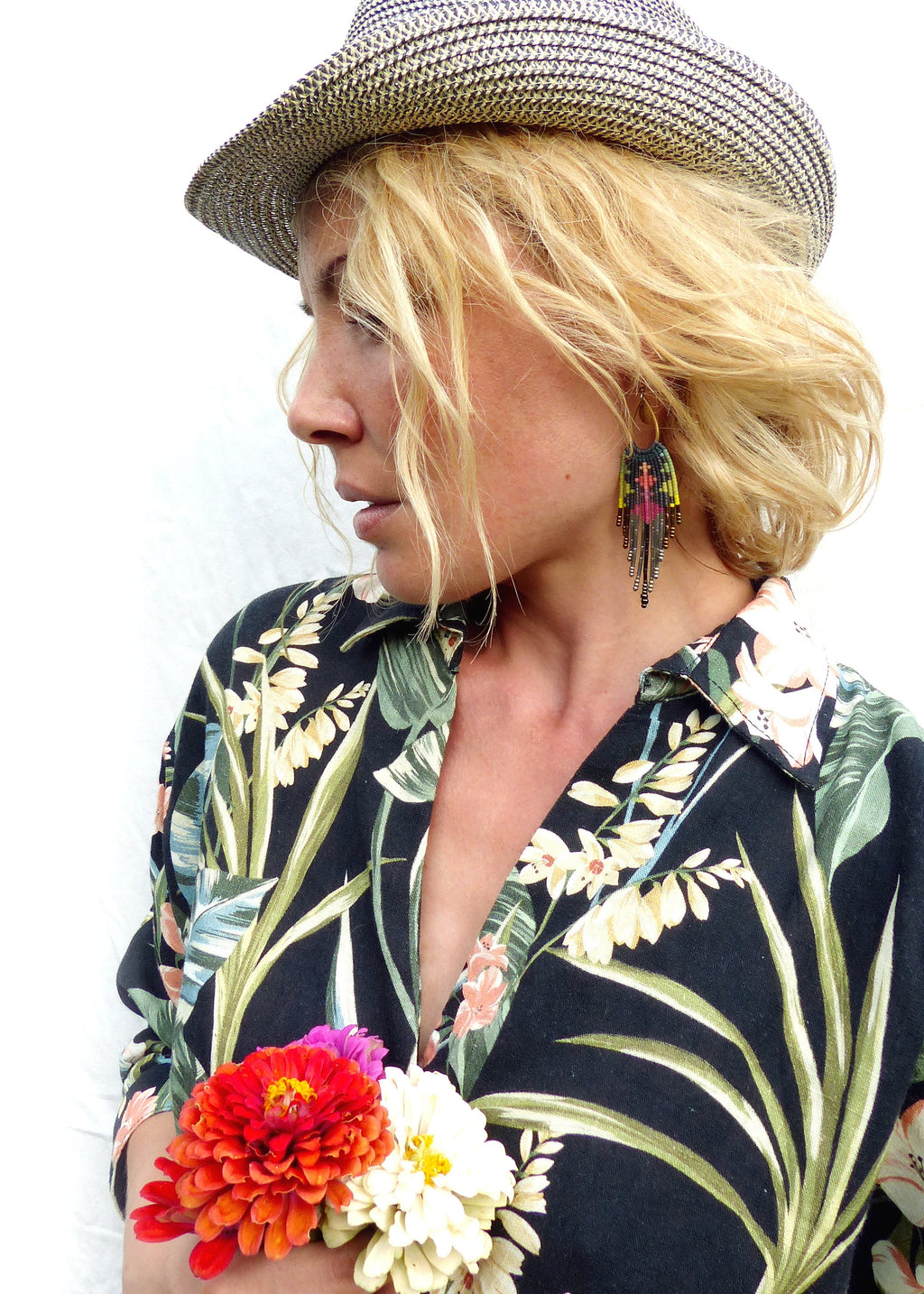Peut inclure: Une femme portant un chapeau de paille et une chemise à fleurs noire et verte à manches longues. Elle tient un bouquet de fleurs roses et orange. Elle porte de longues boucles d'oreilles en perles avec un motif coloré.