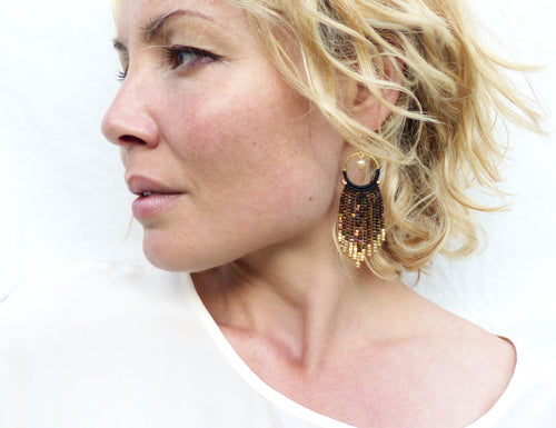 Peut inclure: Une paire de boucles d'oreilles en perles dorées et noires avec un design en frange. Les boucles d'oreilles pendent des lobes d'oreille d'une femme aux cheveux blonds.