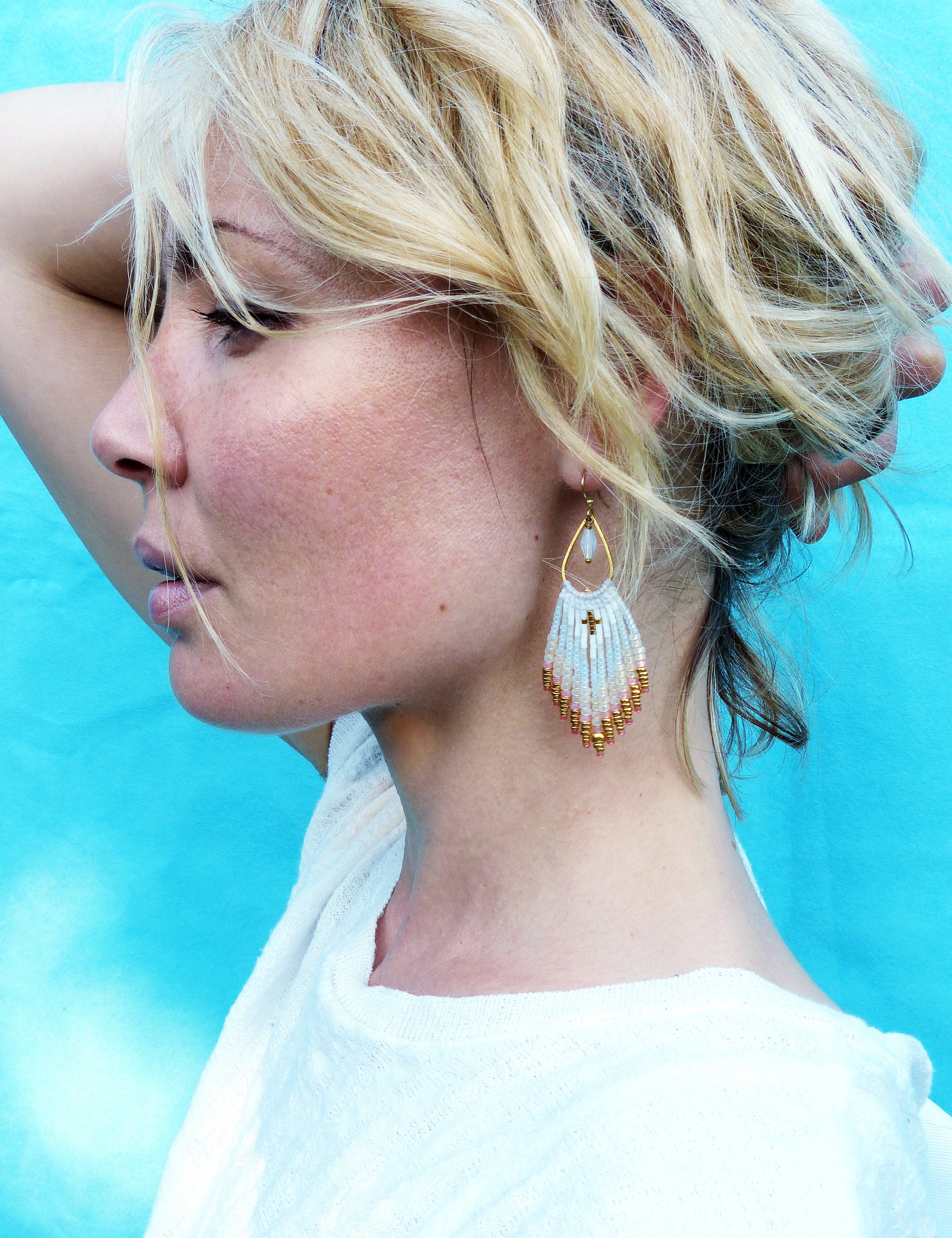 Peut inclure: Une femme aux cheveux blonds portant une chemise blanche et des boucles d'oreilles en perles avec un pendentif en forme de croix dorée. Les boucles d'oreilles sont blanches et dorées et ont une forme de larme.