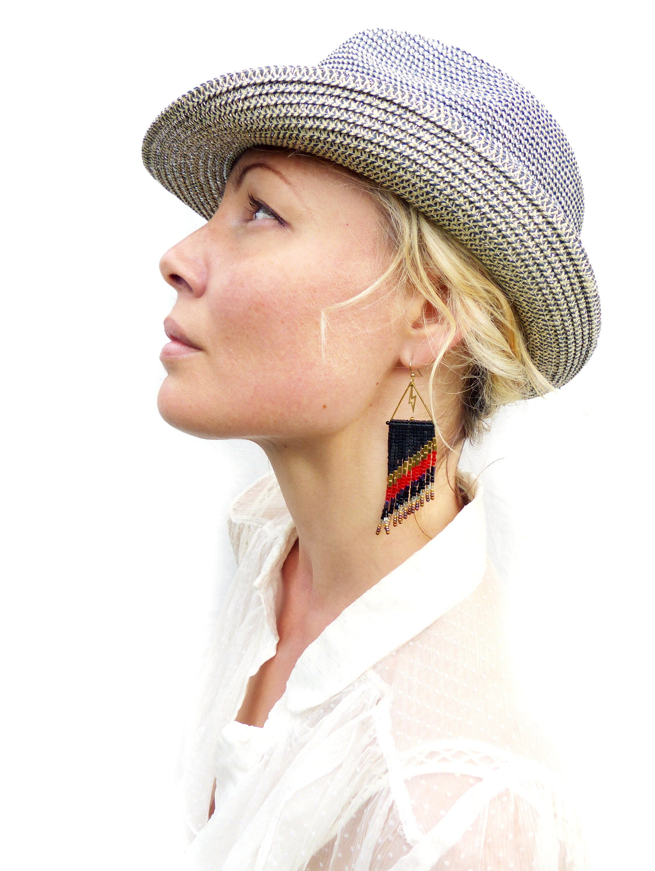 Peut inclure: Une femme porte un chapeau de paille avec un motif tissé noir et blanc et des boucles d'oreilles dorées. Les boucles d'oreilles sont triangulaires avec un motif de perles noir, rouge et or. La femme porte un chemisier blanc.