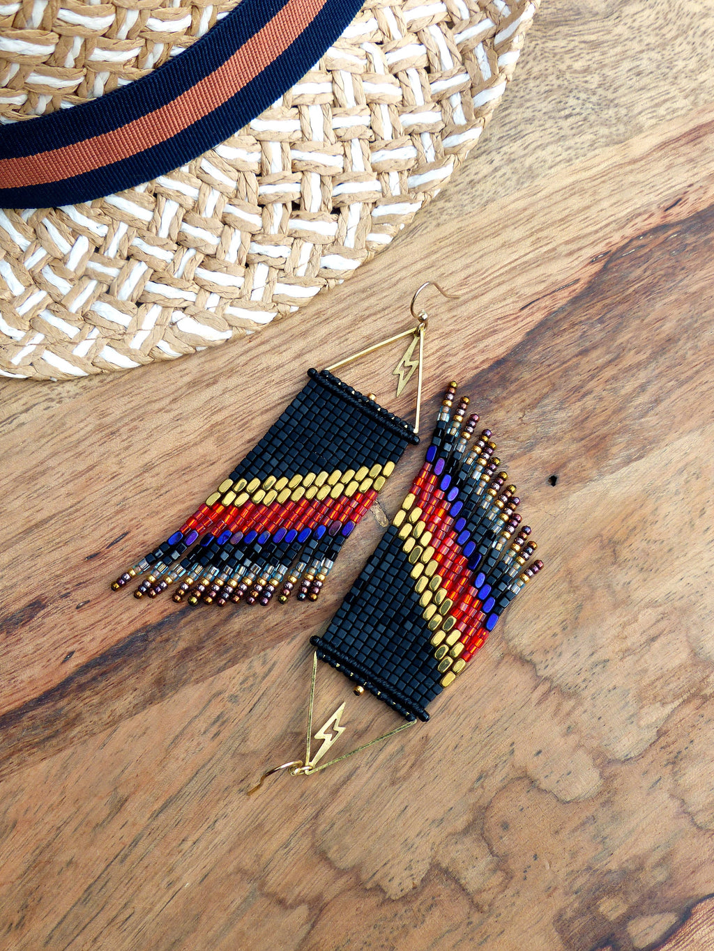 Peut inclure: Boucles d'oreilles en perles avec une base noire et des rayures colorées rouges, jaunes et bleues. Elles présentent un motif d'éclair doré et sont posées sur une surface en bois, avec un chapeau de paille en arrière-plan.