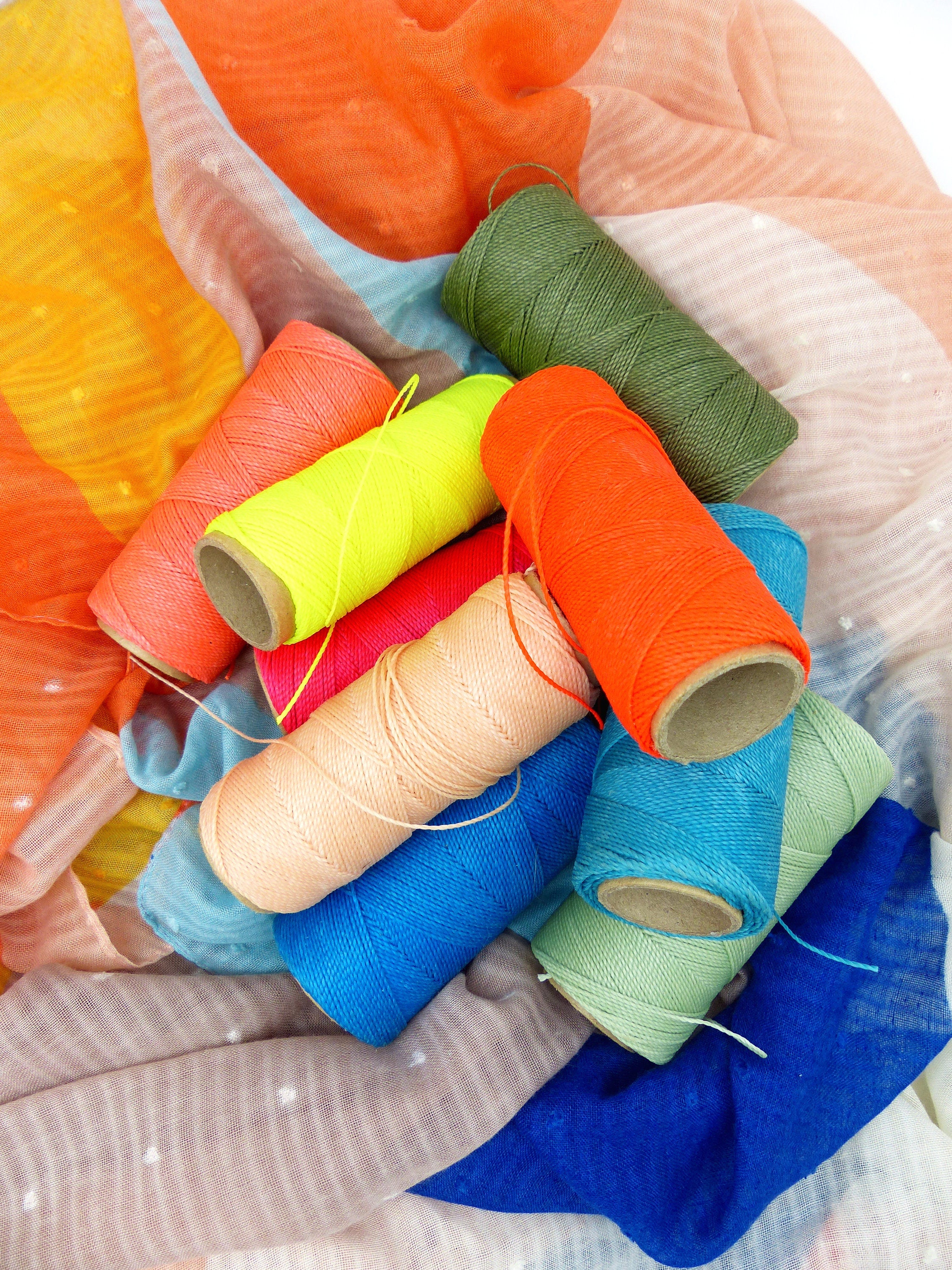 Peut inclure: Plusieurs bobines de fil colorées, aux teintes vives d'orange, de vert, de bleu, de rose et de jaune. Les bobines sont disposées sur un tissu à motifs aux tons orange, bleu et blanc. L'image est bien éclairée, mettant en valeur les textures et les couleurs.