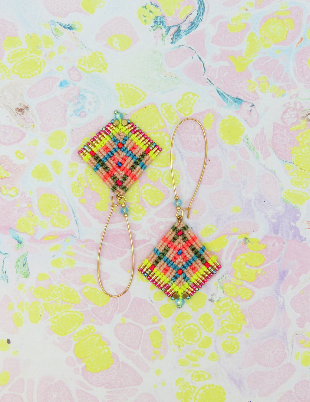 Peut inclure: Une paire de boucles d'oreilles en macramé faites à la main. Chaque boucle d'oreille présente un motif en forme de losange avec un motif coloré de fils roses, verts, jaunes et bleus. Les boucles d'oreilles ont des crochets dorés et de petites perles bleues.
