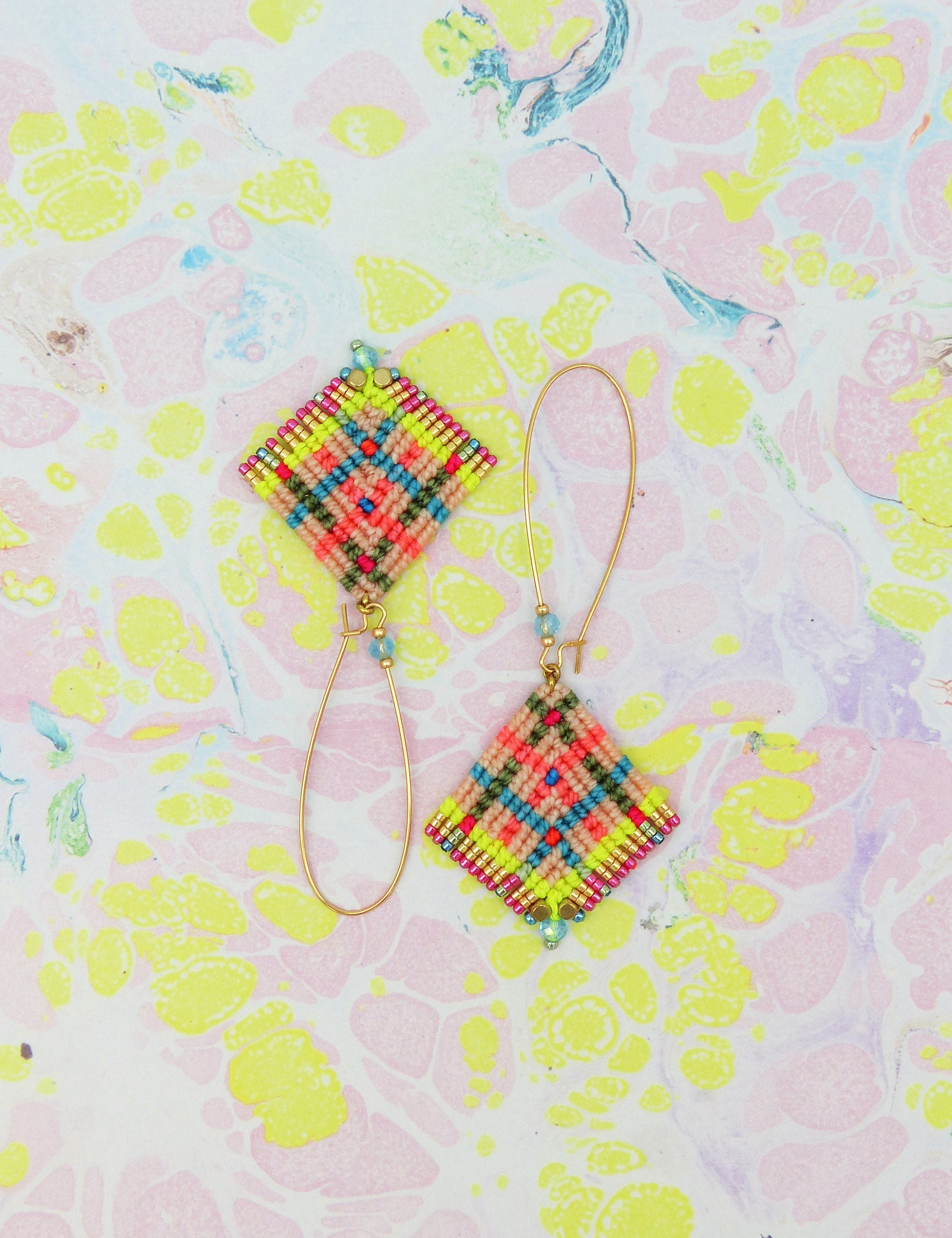 Peut inclure: Une paire de boucles d'oreilles en macramé faites à la main. Chaque boucle d'oreille présente un motif en forme de losange avec un motif coloré de fils roses, verts, jaunes et bleus. Les boucles d'oreilles ont des crochets dorés et de petites perles bleues.