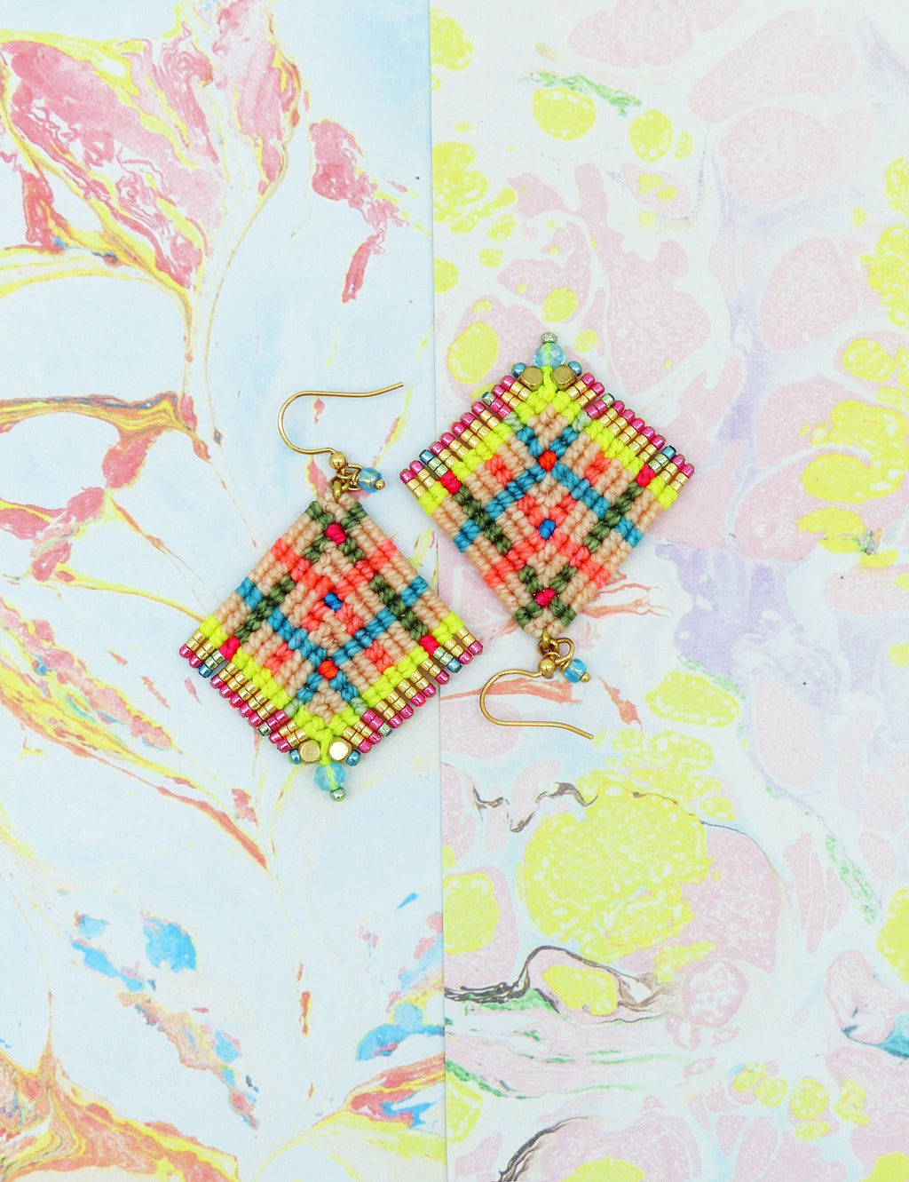 Peut inclure: Une paire de boucles d'oreilles en forme de losange aux couleurs vives. Elles présentent un motif tissé en jaune fluo, corail, turquoise et beige. Des crochets dorés et de petites perles bleues complètent le design. Le fond est un motif marbré aux couleurs pastel.