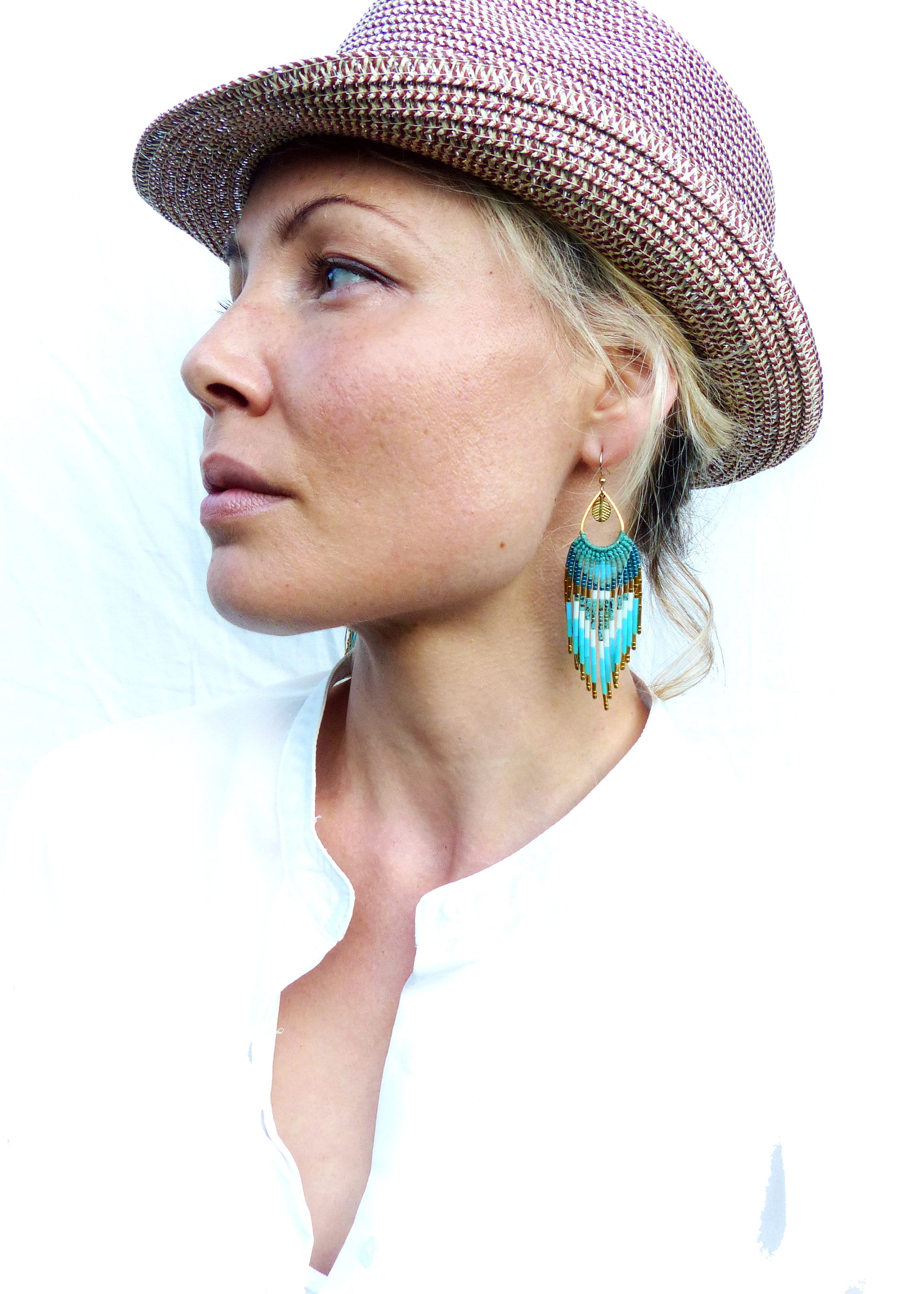 Peut inclure: Boucles d'oreilles dorées avec des franges de perles turquoise, blanches et marron. Les boucles d'oreilles ont une forme de goutte d'eau avec un accent de feuille d'or. La personne porte une chemise blanche et un chapeau marron et beige.