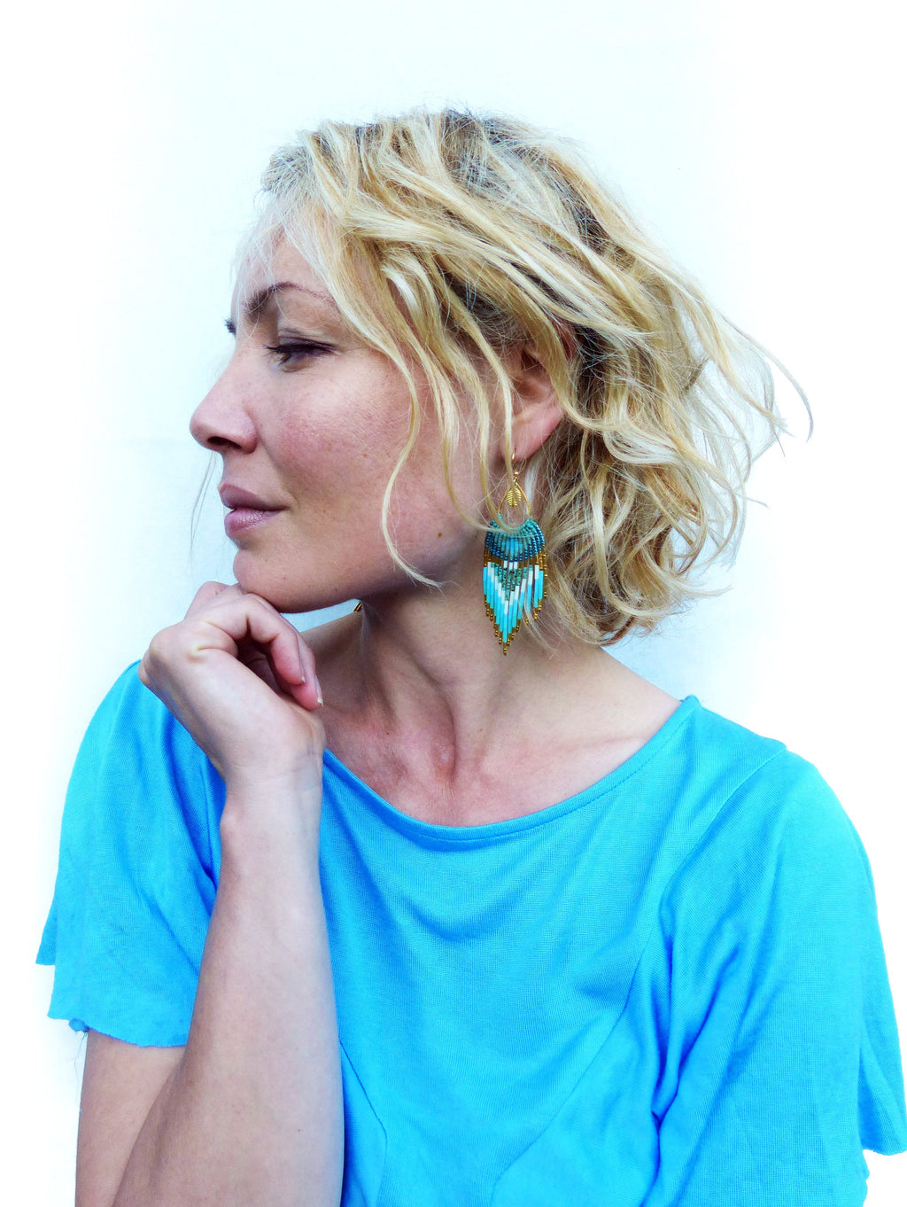 Peut inclure: Une femme porte des boucles d'oreilles en perles turquoise et dorées. Les boucles d'oreilles ont un motif géométrique avec des perles turquoise, blanches et dorées. La femme porte un haut bleu turquoise et a les cheveux blonds.