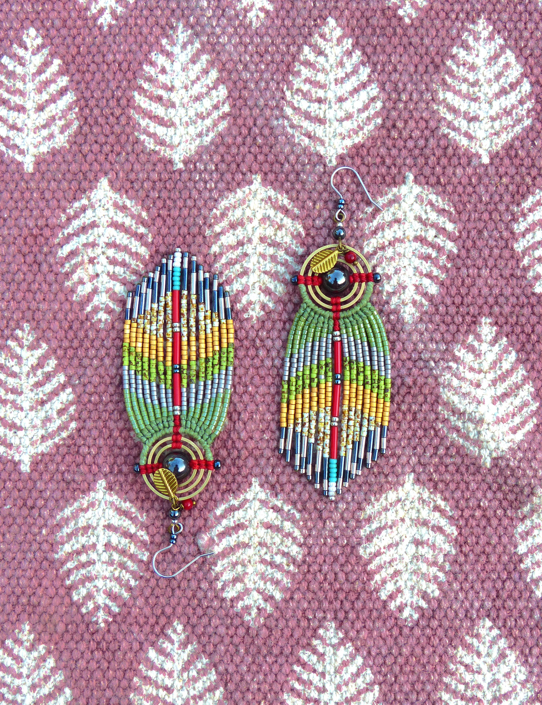Peut inclure: Une paire de longues boucles d'oreilles en perles avec un motif géométrique. Les boucles d'oreilles sont faites de perles vertes, argentées, rouges et dorées. Les boucles d'oreilles ont un motif circulaire au centre avec une pierre sombre et une feuille d'or.