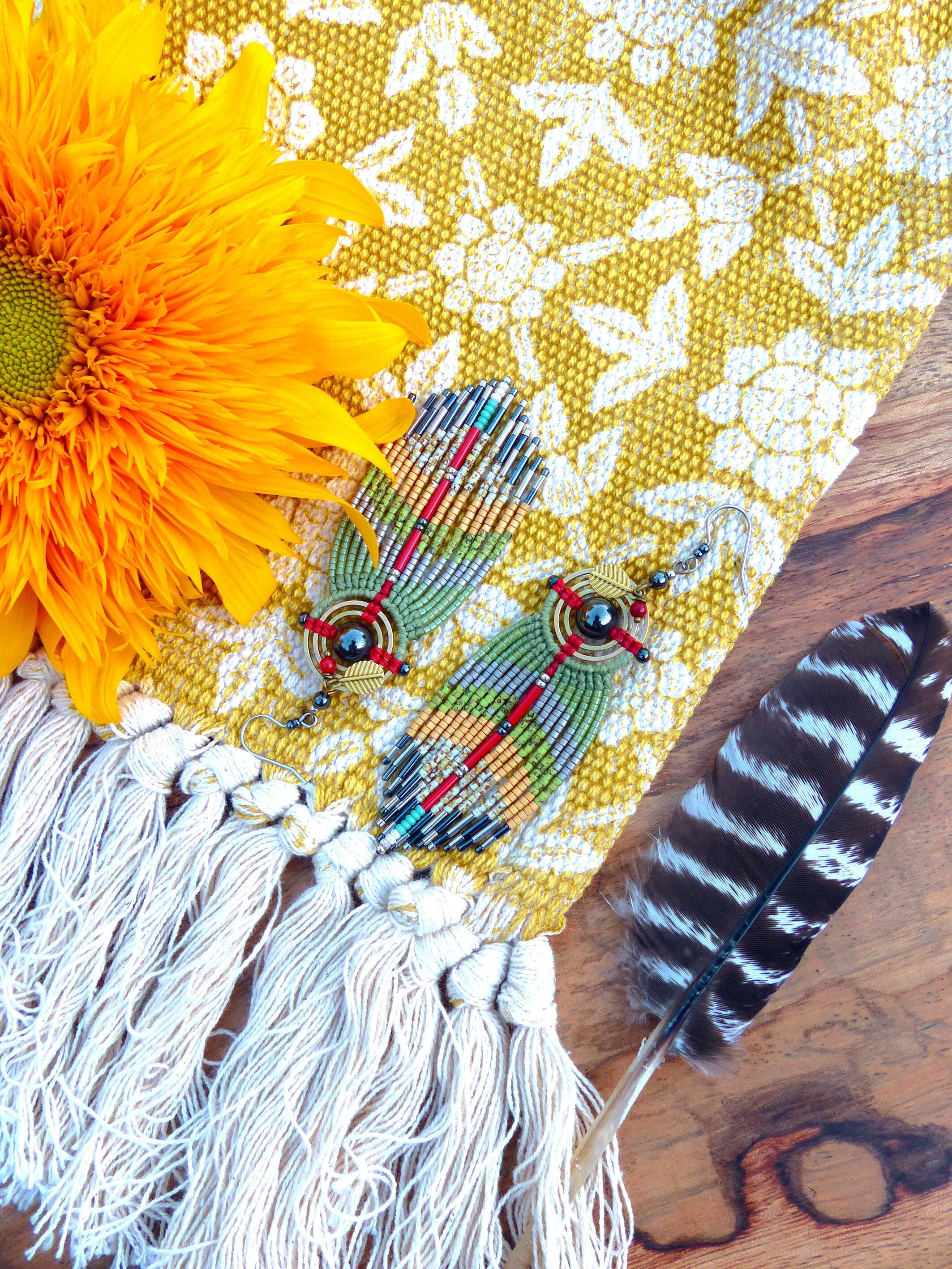Peut inclure: Une paire de boucles d'oreilles en perles avec un schéma de couleurs vert, jaune et rouge. Les boucles d'oreilles sont suspendues à un tissu jaune avec un motif floral blanc. Une plume brune et blanche est également dans l'image.