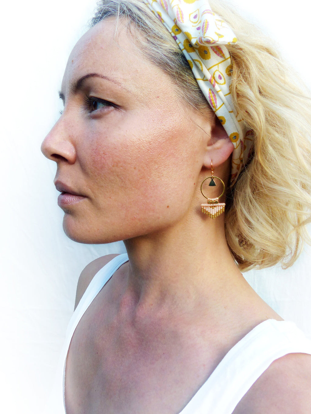 Peut inclure: Une femme portant un débardeur blanc et un bandeau floral avec des boucles d'oreilles en pêche et or. Les boucles d'oreilles ont un petit charme triangulaire et un cercle avec une frange.