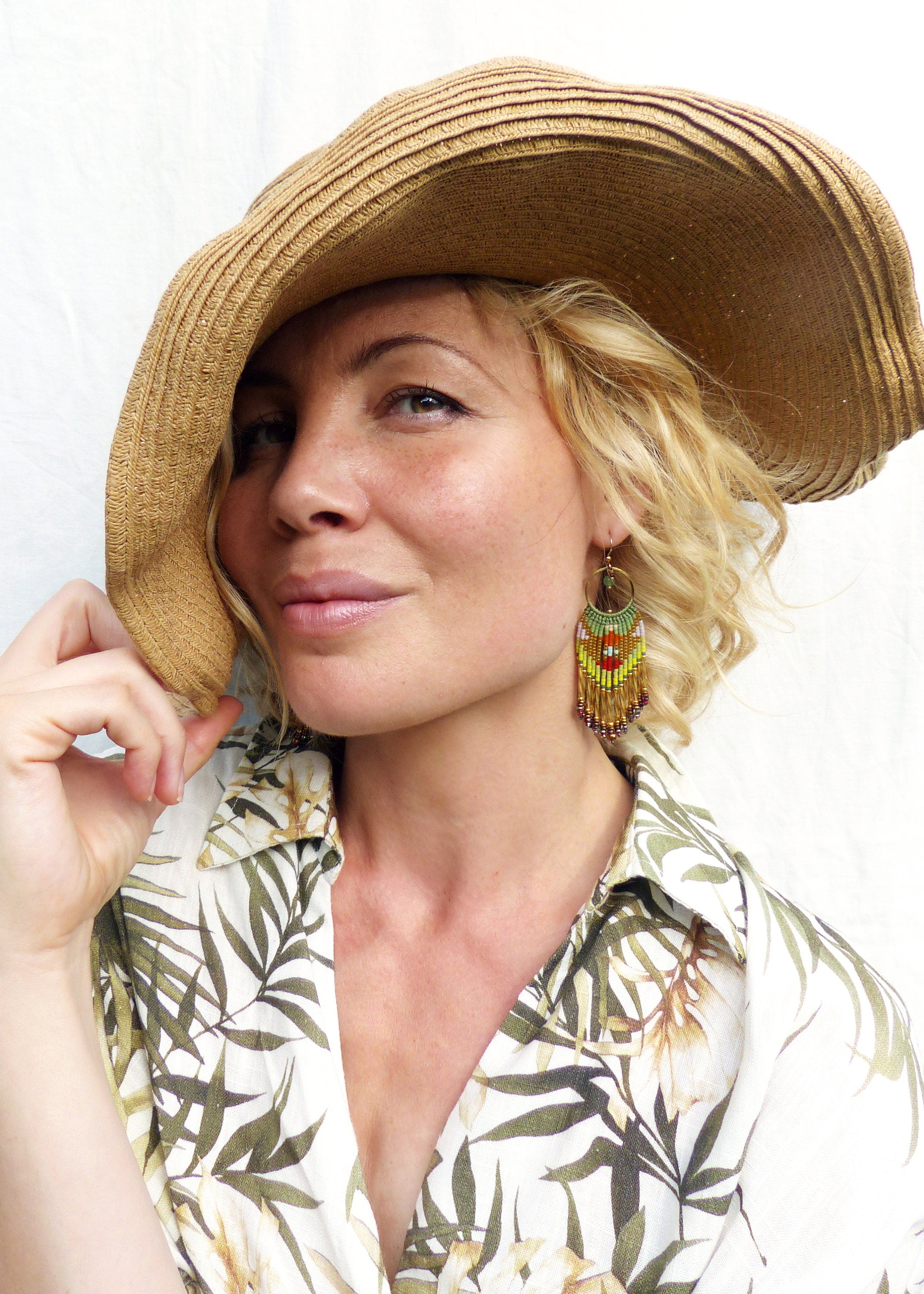 Peut inclure: Une femme portant un chapeau de paille et une chemise blanche à imprimé tropical. Elle porte de longues boucles d'oreilles en perles avec des perles vertes, jaunes, orange et rouges.