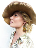 Peut inclure: Une femme portant un chapeau de paille et une chemise blanche à motif floral vert et marron. Elle porte de longues boucles d'oreilles en perles avec des perles vertes, jaunes, rouges et brunes.