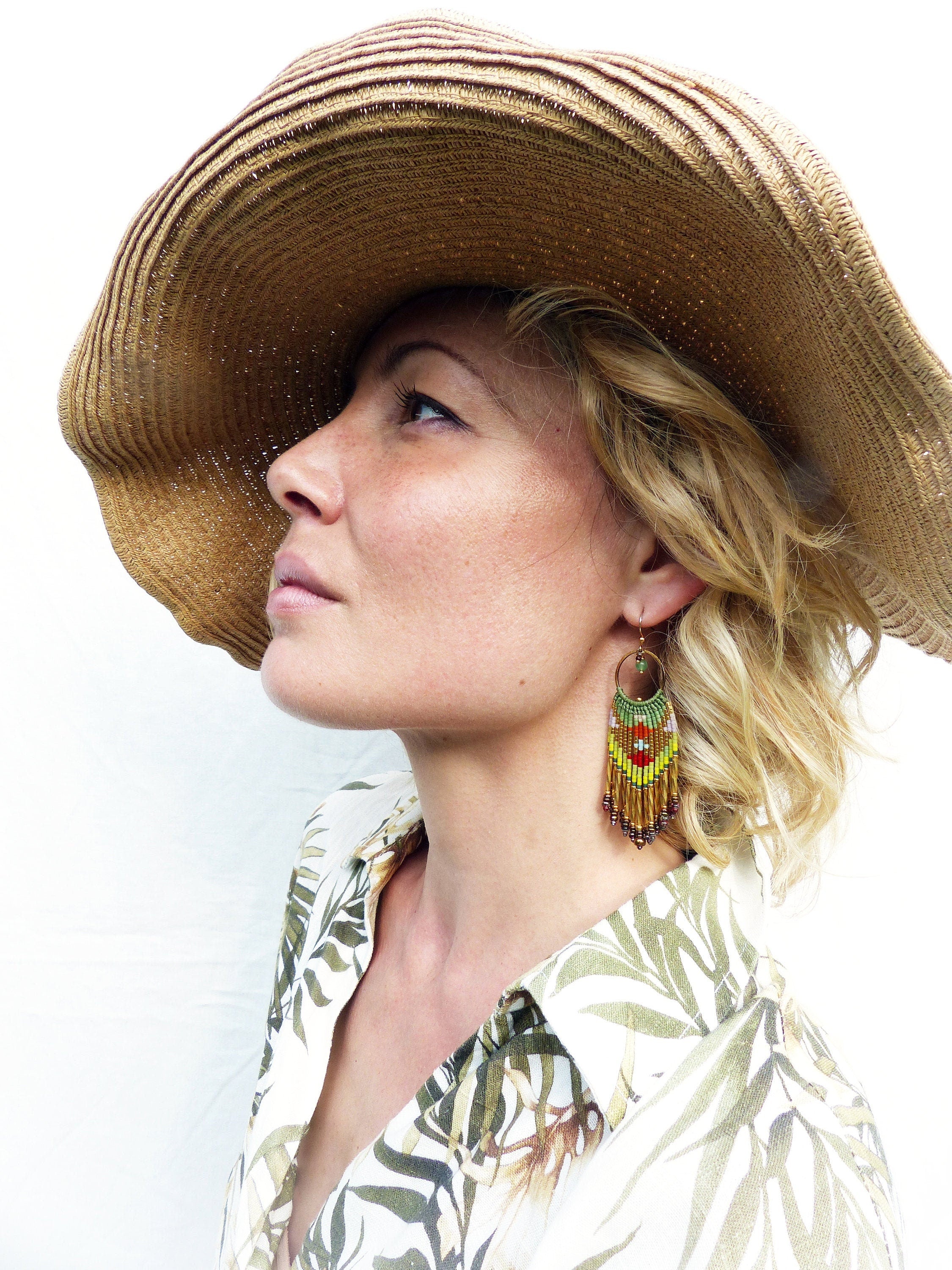 Peut inclure: Une femme portant un chapeau de paille et une chemise blanche à motif floral vert et marron. Elle porte de longues boucles d'oreilles en perles avec des perles vertes, jaunes, rouges et brunes.