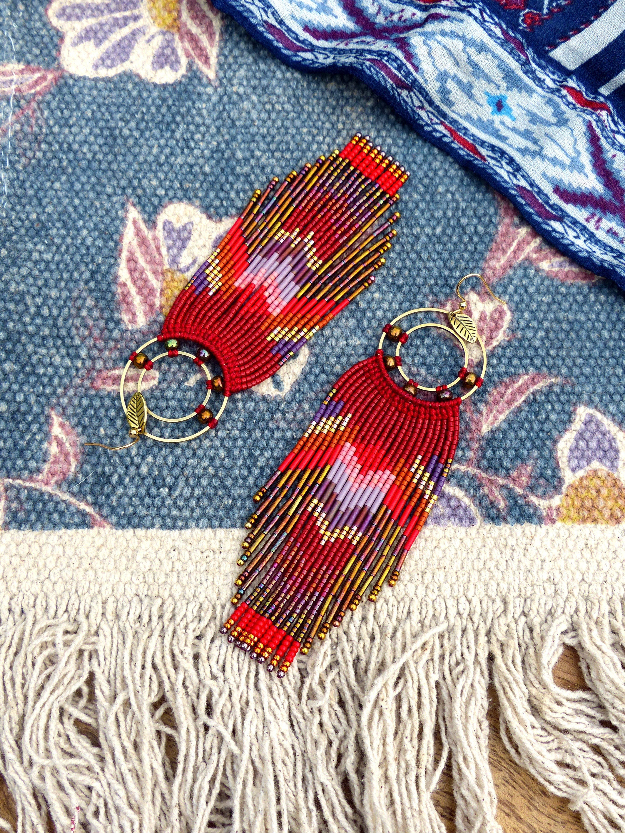 Peut inclure: Une paire de longues boucles d'oreilles rouges en perles avec des cercles dorés et un charme en forme de feuille. Les boucles d'oreilles sont faites avec une variété de perles rouges, dorées et violettes.