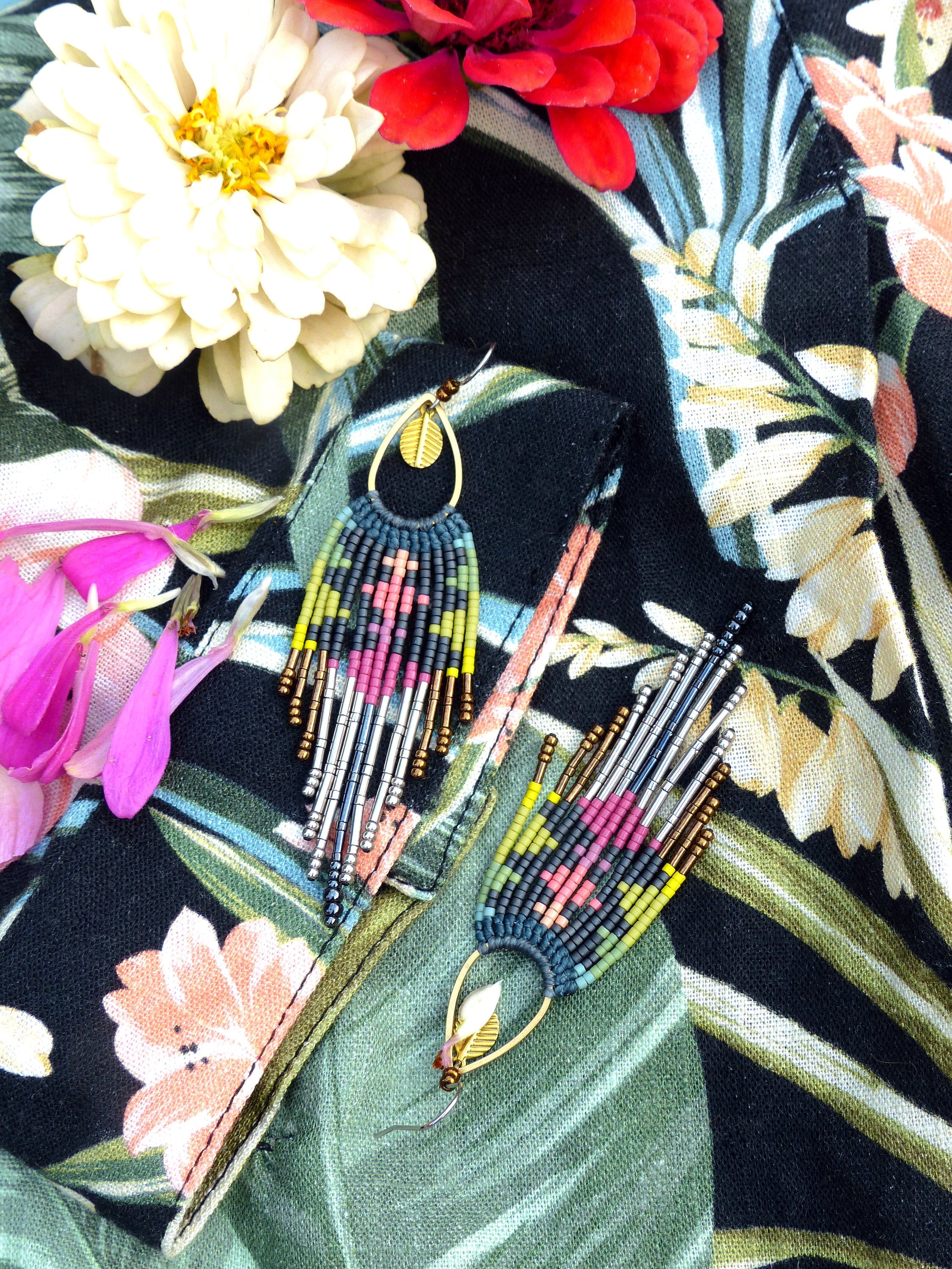 Peut inclure: Une paire de longues boucles d'oreilles en perles avec un charme en feuille d'or. Les boucles d'oreilles sont faites de différentes couleurs, dont le rose, le vert, le bleu et le noir. Les boucles d'oreilles sont suspendues à un tissu noir à motif floral.