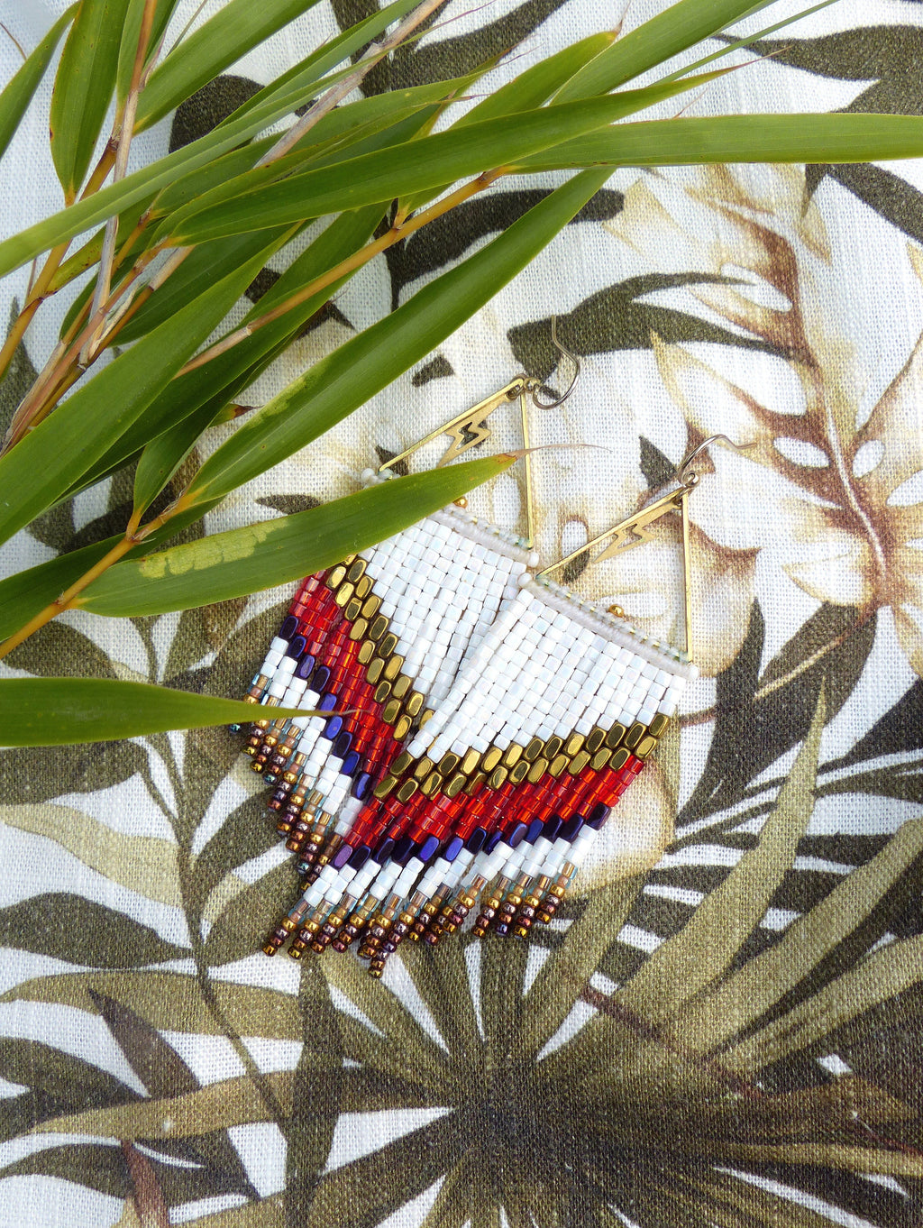 Peut inclure: Boucles d'oreilles en perles blanches, rouges et dorées avec un motif géométrique. Les boucles d'oreilles sont suspendues à un crochet en métal doré.