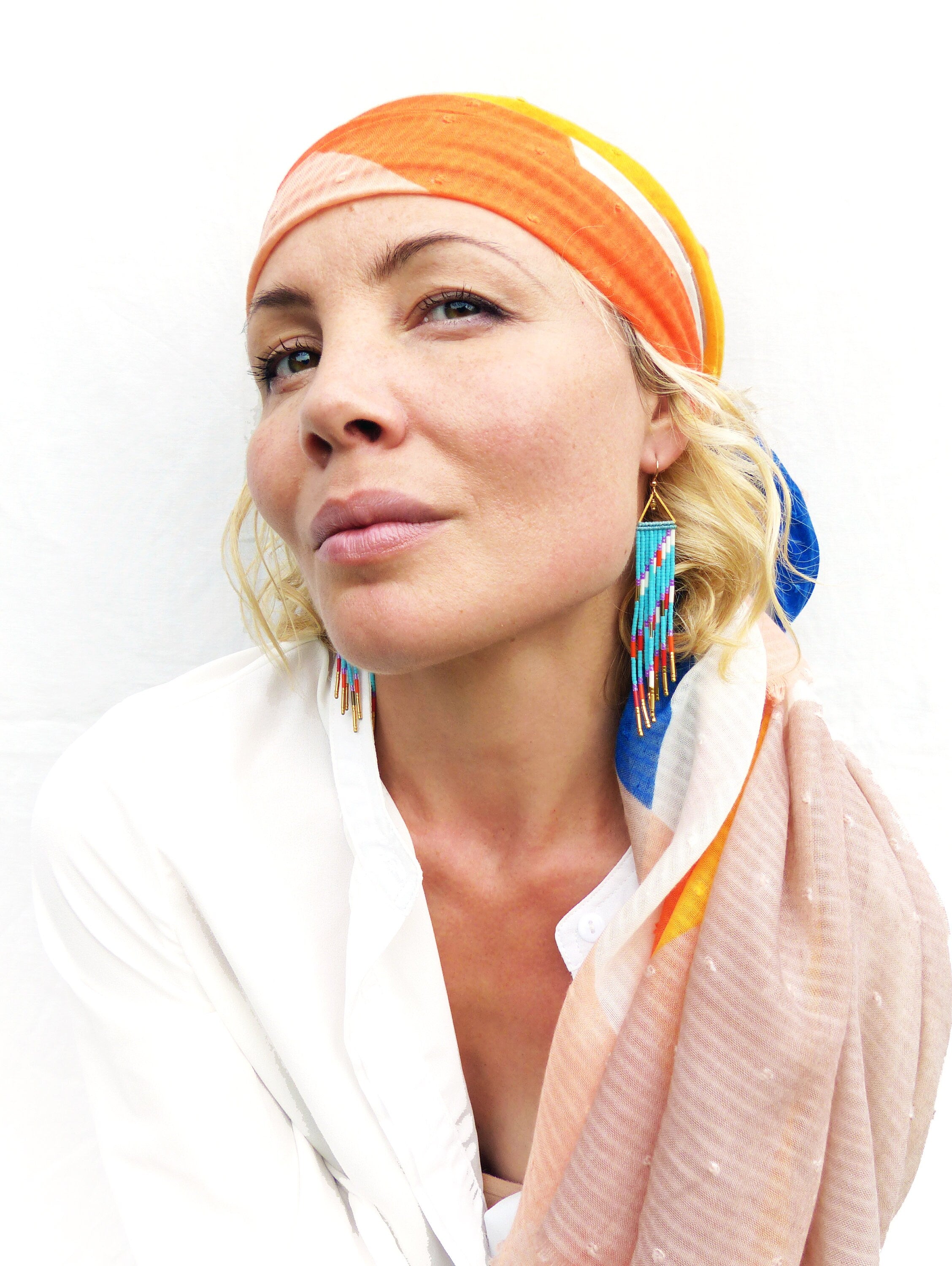 Peut inclure: Une femme portant une chemise blanche et une écharpe colorée à motif géométrique. Elle porte de longues boucles d'oreilles en perles turquoise avec des accents dorés.