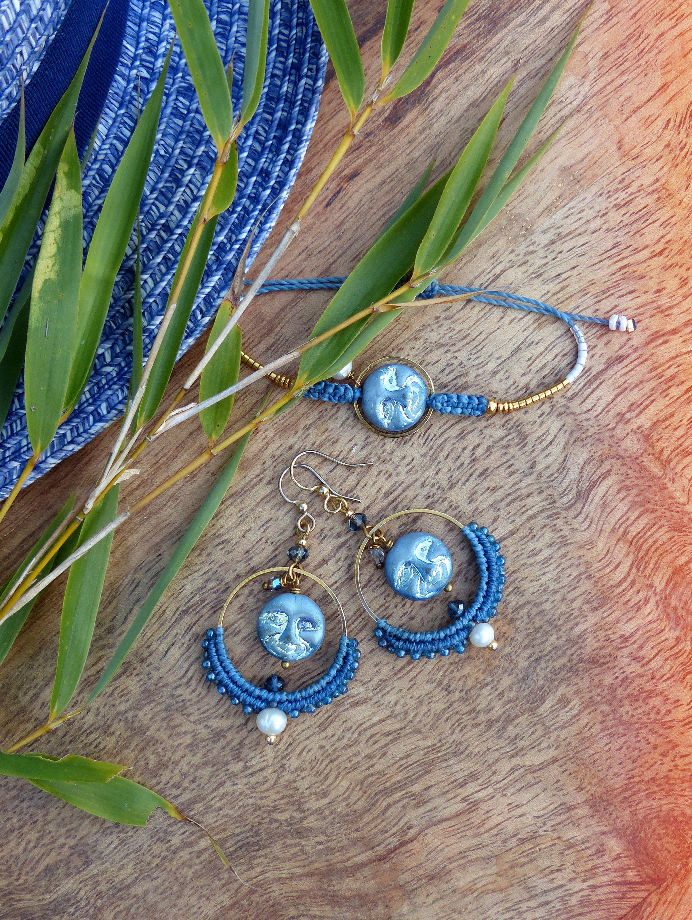 Peut inclure: Un ensemble de bijoux bleu et or avec un pendentif en forme de lune et des boucles d'oreilles. Les boucles d'oreilles sont faites de perles bleues et d'un accent de perle. Les bijoux sont présentés sur une surface en bois avec des feuilles vertes.