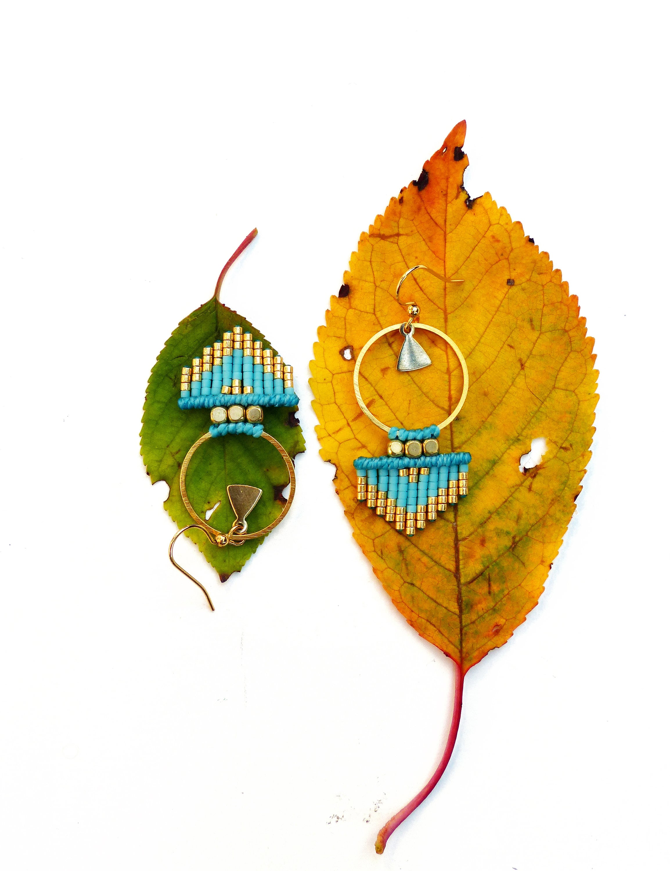 Peut inclure: Une paire de boucles d'oreilles en perles turquoise et dorées avec un motif géométrique. Les boucles d'oreilles sont suspendues à une feuille sur un fond blanc.