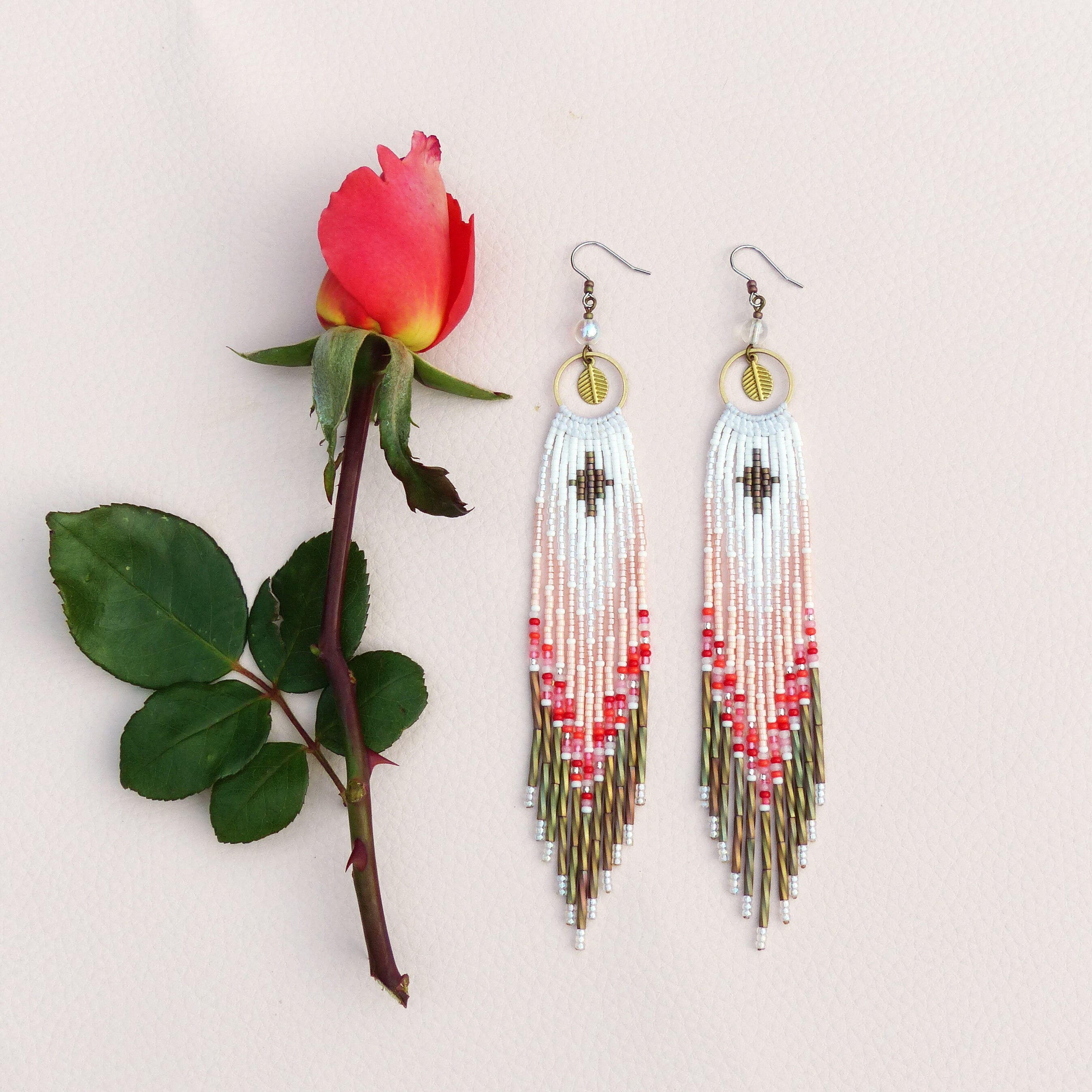 Peut inclure: Une paire de longues boucles d'oreilles en perles avec un motif blanc, rose et rouge. Les boucles d'oreilles présentent un charme en forme de feuille dorée et un motif géométrique.