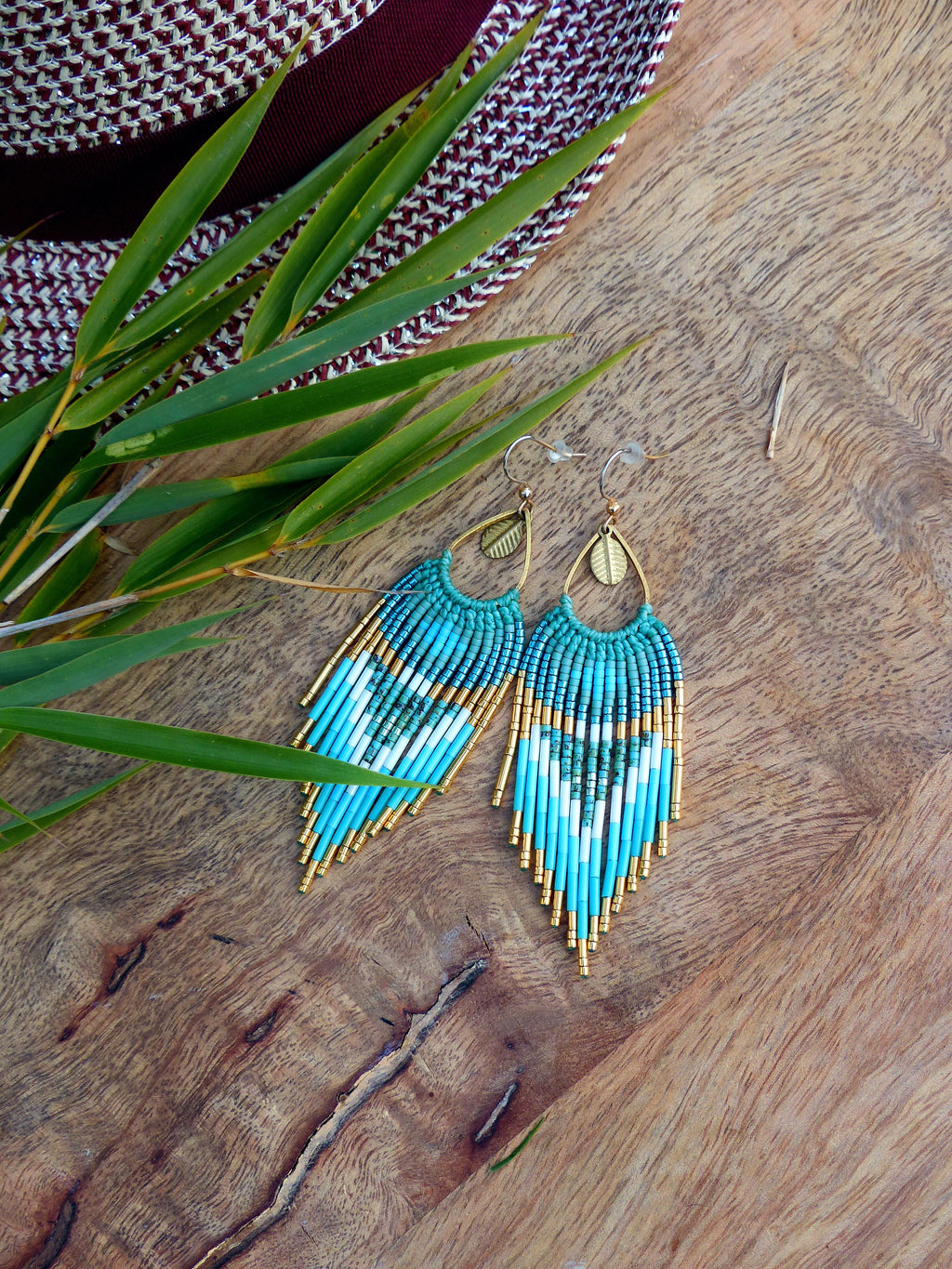 Peut inclure: Une paire de boucles d'oreilles turquoise perlées avec des accents dorés et des détails en forme de feuille. Les boucles d'oreilles présentent un motif géométrique avec des perles blanches et dorées, et sont suspendues à des crochets d'oreilles dorés.