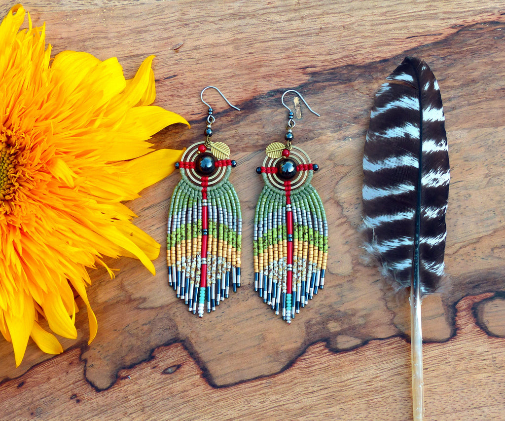Peut inclure: Une paire de longues boucles d'oreilles en perles avec un schéma de couleurs vert, rouge et or. Les boucles d'oreilles présentent un motif circulaire avec une pierre noire au centre et un petit accent en feuille d'or. Les boucles d'oreilles sont suspendues sur un fond en bois.