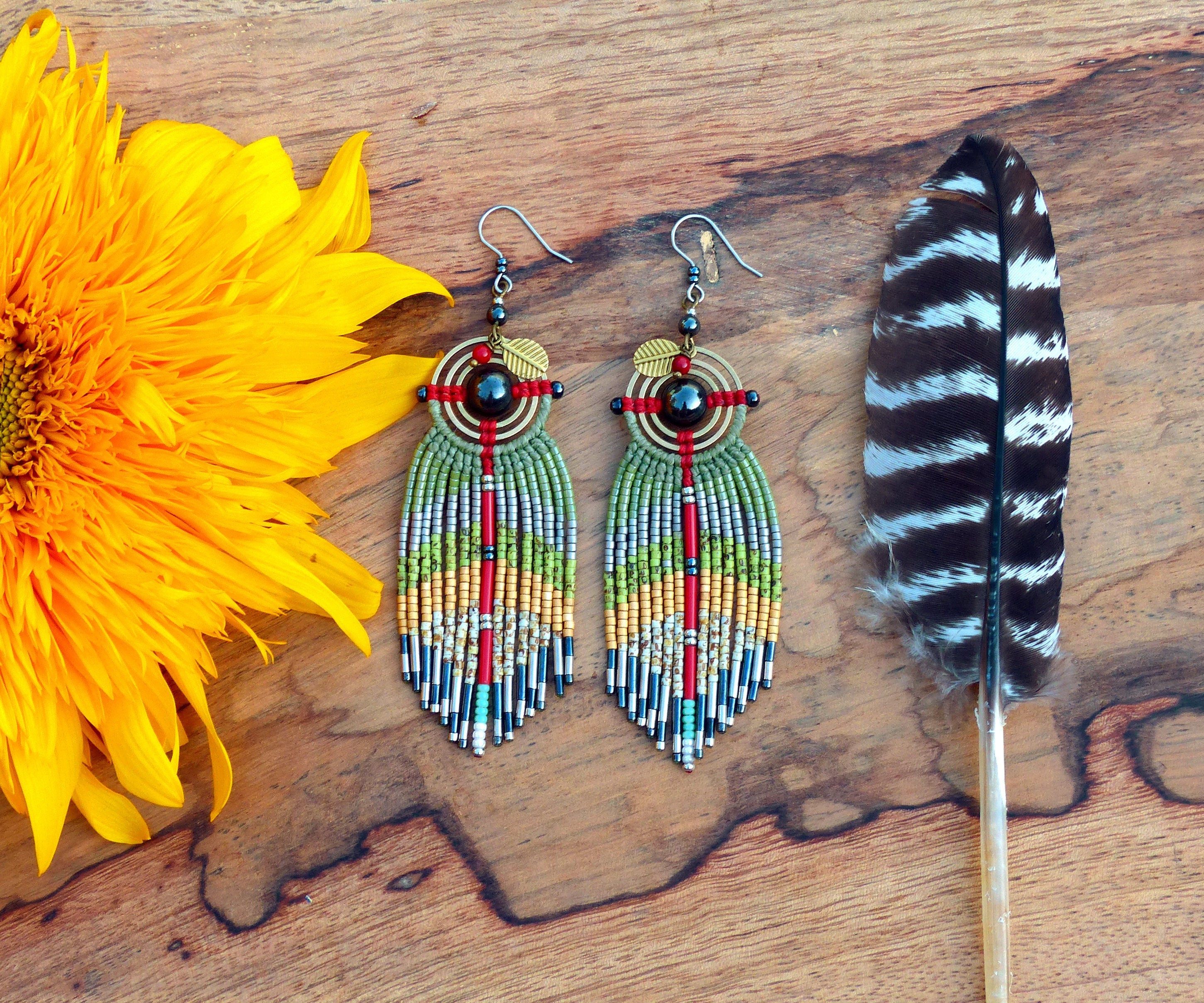 Peut inclure: Une paire de longues boucles d'oreilles en perles avec un schéma de couleurs vert, rouge et or. Les boucles d'oreilles présentent un motif circulaire avec une pierre noire au centre et un petit accent en feuille d'or. Les boucles d'oreilles sont suspendues sur un fond en bois.