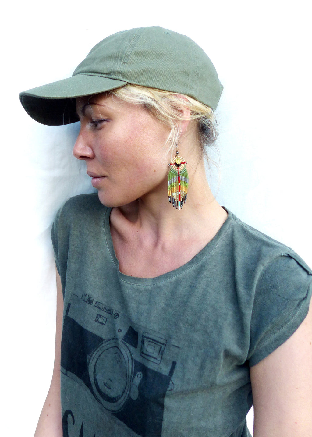 Peut inclure: Une femme portant une casquette de baseball verte et un t-shirt vert avec un motif de caméra noir. Elle porte de longues boucles d'oreilles en perles colorées.