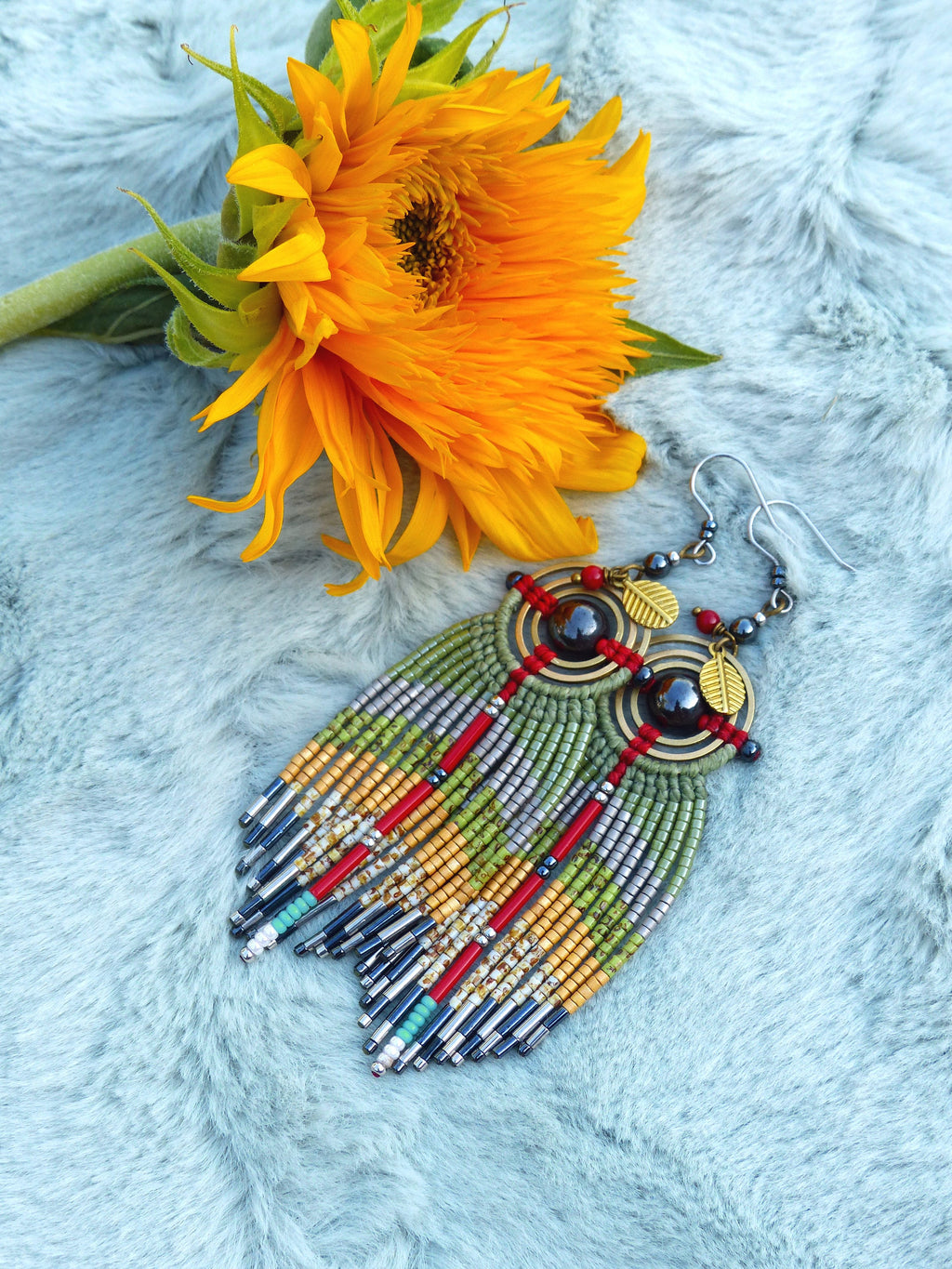 Peut inclure: Une paire de longues boucles d'oreilles en perles avec un schéma de couleurs vert, rouge et or. Les boucles d'oreilles présentent un pendentif rond en perles avec une pierre sombre et des accents dorés. Les boucles d'oreilles sont présentées sur une surface floue bleu clair avec un tournesol jaune.