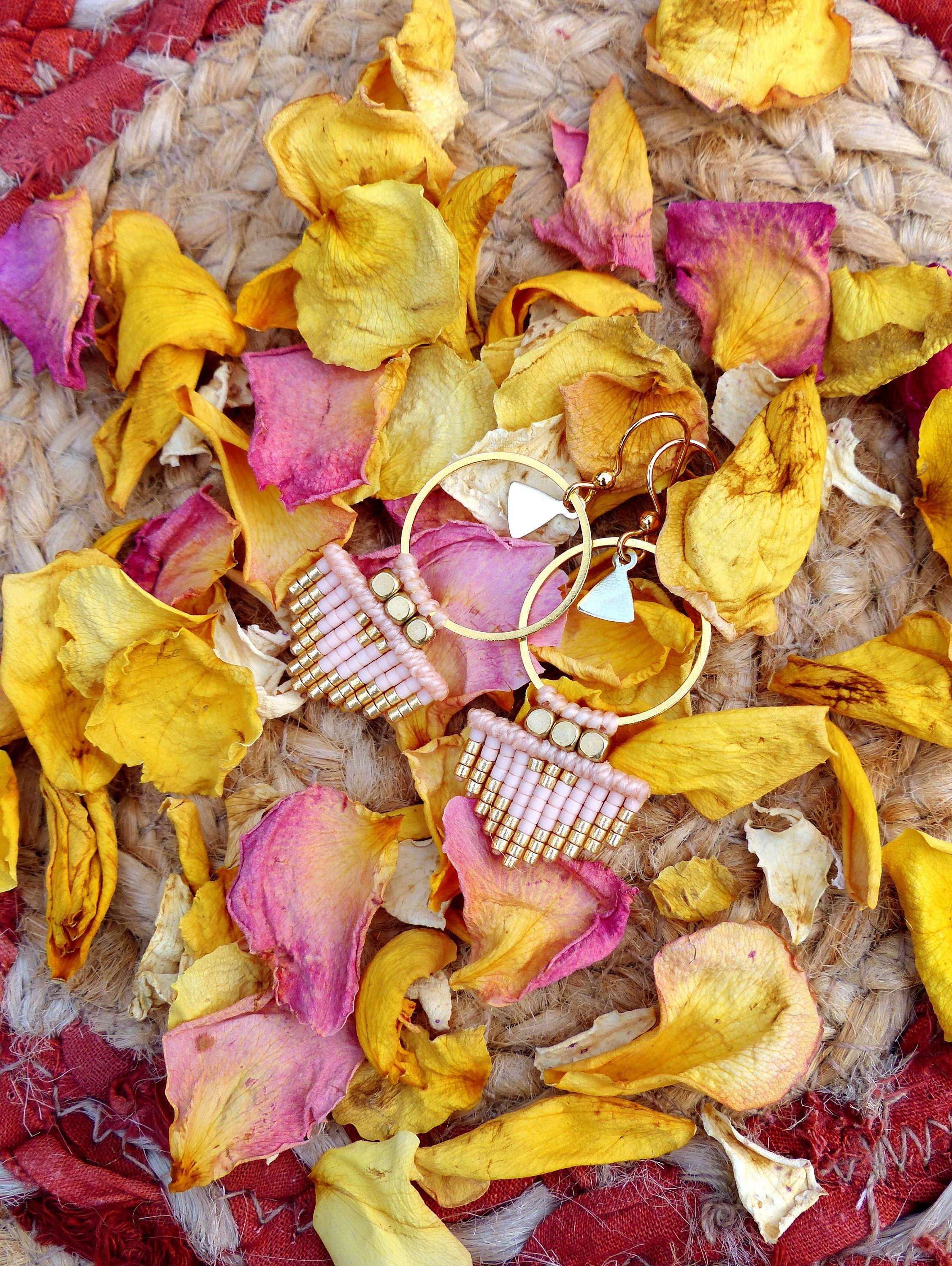 Peut inclure: Une paire de boucles d'oreilles créoles dorées avec des franges de perles roses et dorées qui pendent du bas. Les boucles d'oreilles sont photographiées sur un fond de pétales de roses séchées.