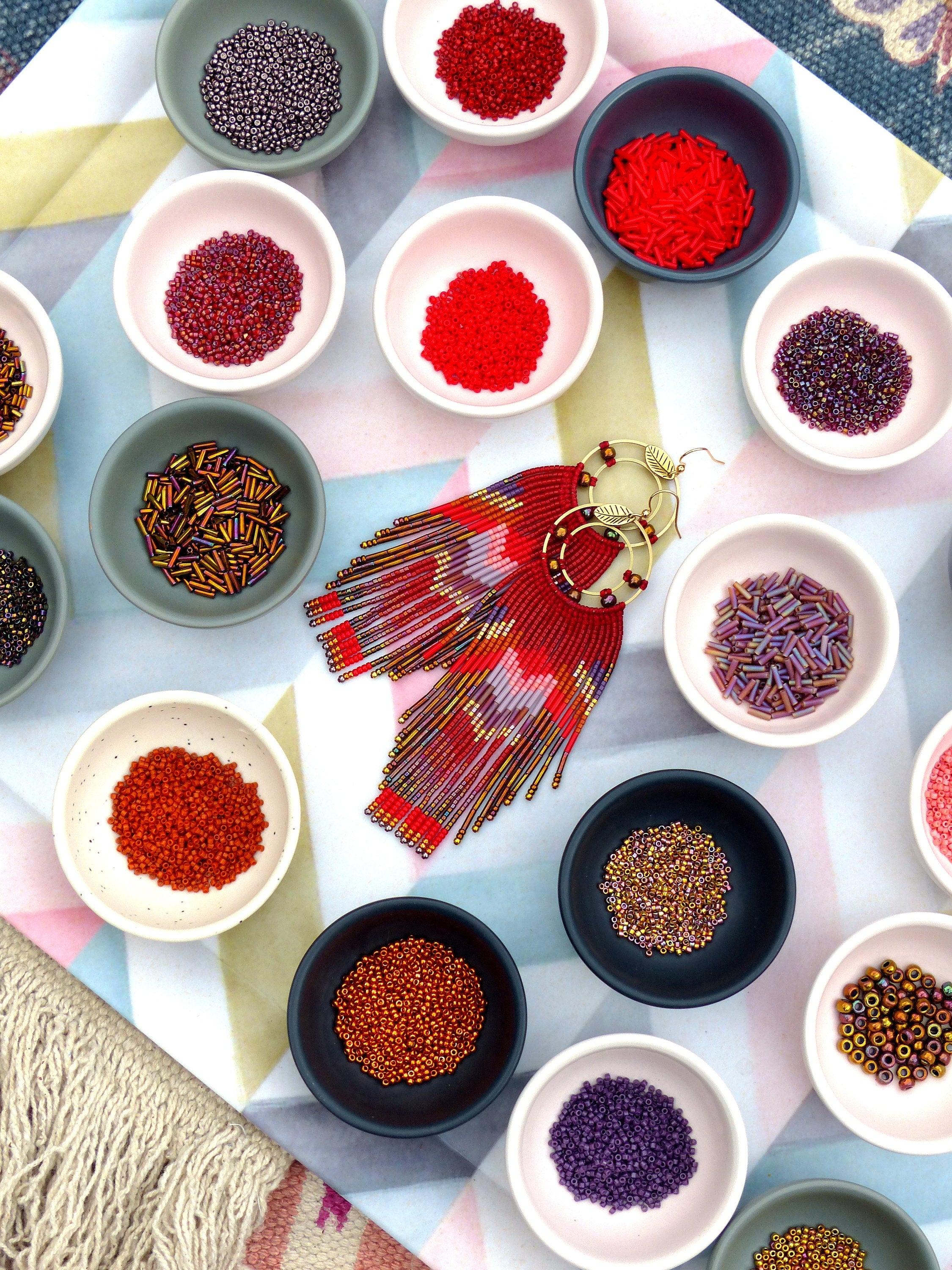 Peut inclure: Un assortiment de perles colorées dans de petits bols, y compris du rouge, de l'orange, du violet et de l'or. Une paire de boucles d'oreilles en perles avec des perles rouges, orange et dorées pend au centre de l'image.