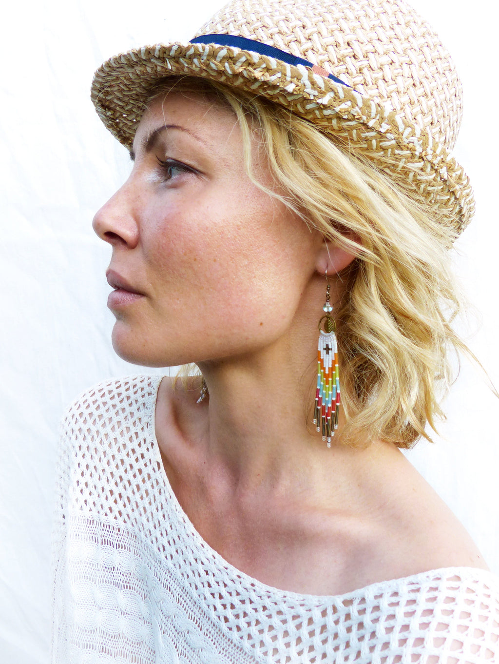 Peut inclure: Une femme portant un chapeau de paille avec un bandeau bleu et un haut en crochet blanc. Elle porte de longues boucles d'oreilles en perles colorées.