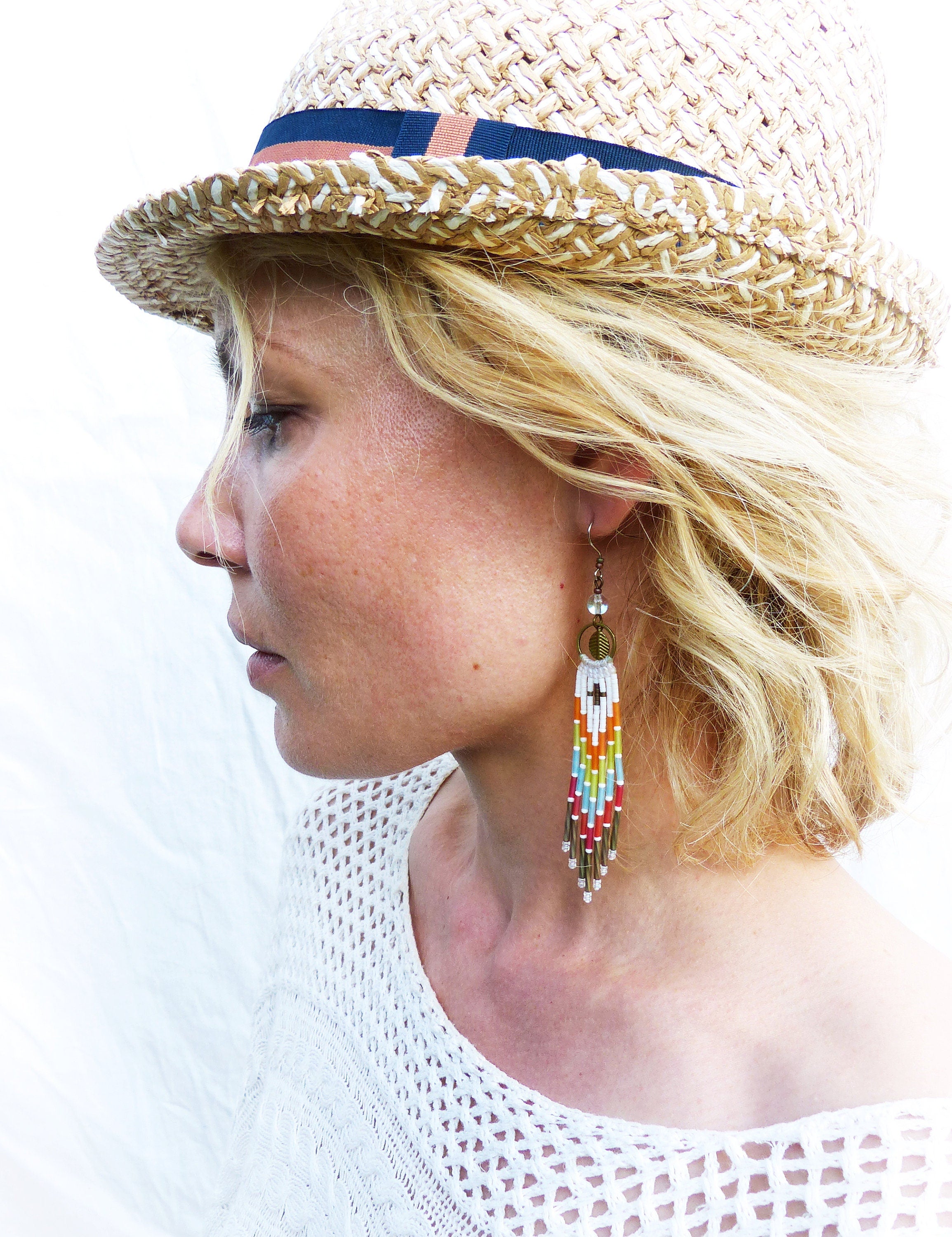 Peut inclure: Une femme portant un chapeau de paille avec un bandeau bleu et un haut en crochet blanc. Elle porte de longues boucles d'oreilles en perles colorées.