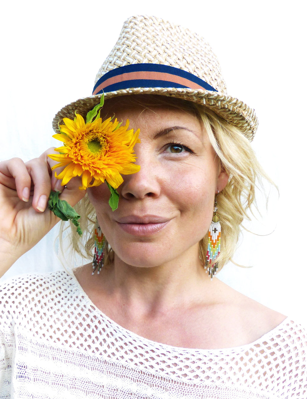 Peut inclure: Une femme portant un chapeau de paille avec un bandeau bleu et un haut en crochet blanc, tenant un tournesol jaune devant son œil. Elle porte des boucles d'oreilles en perles colorées.