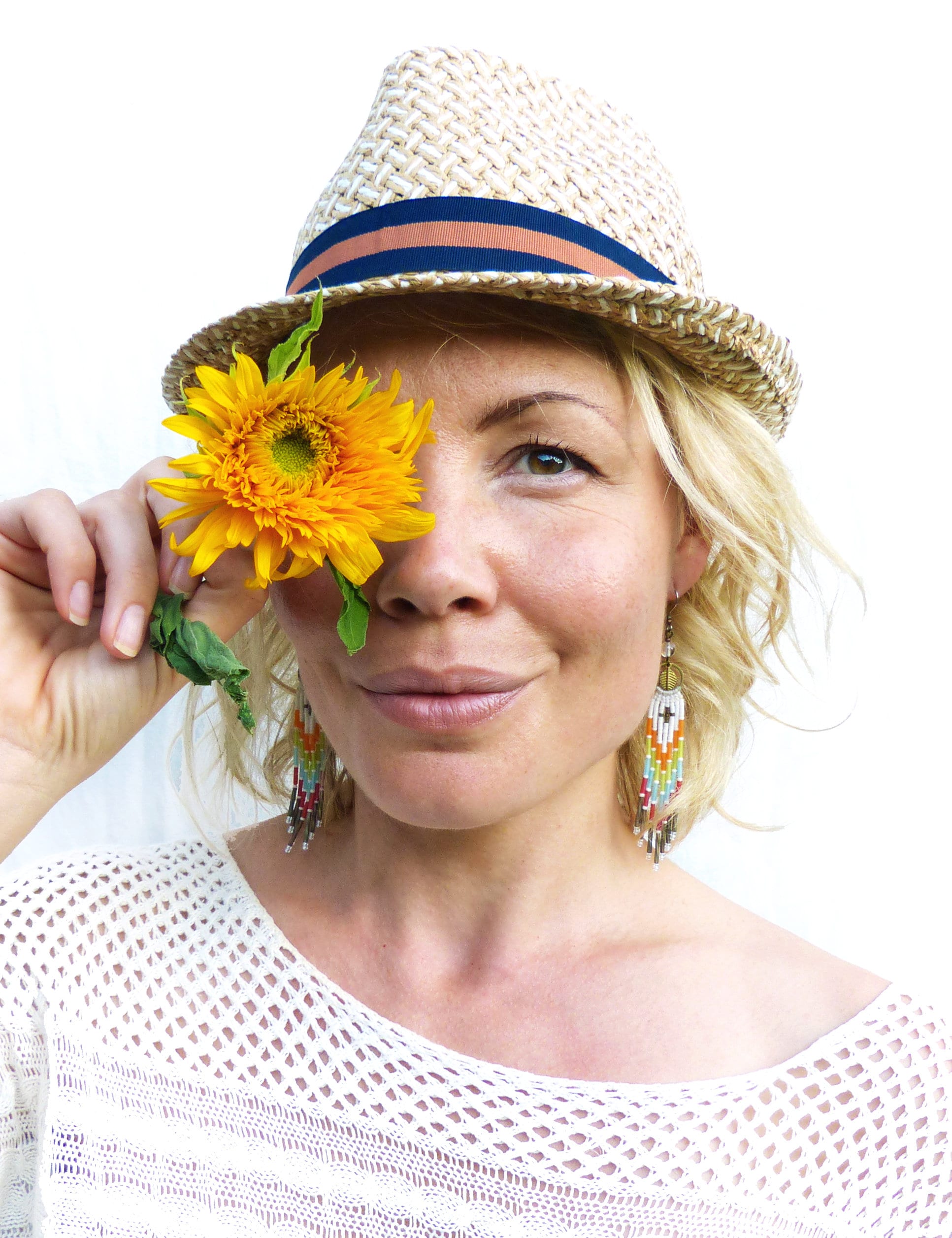 Peut inclure: Une femme portant un chapeau de paille avec un bandeau bleu et un haut en crochet blanc, tenant un tournesol jaune devant son œil. Elle porte des boucles d'oreilles en perles colorées.