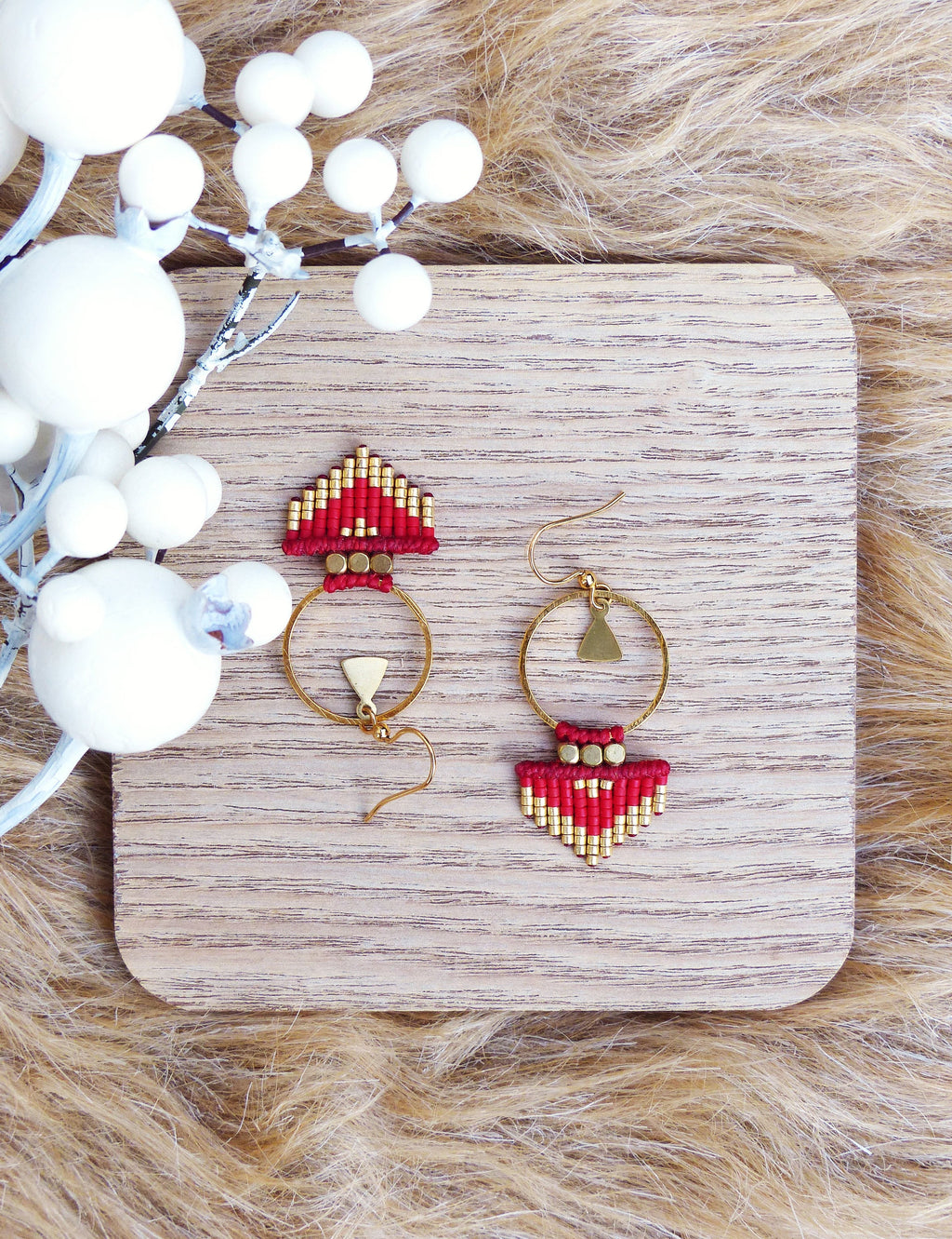 Peut inclure: Une paire de boucles d'oreilles en perles rouges et dorées avec un motif géométrique. Les boucles d'oreilles sont suspendues à une surface en bois.