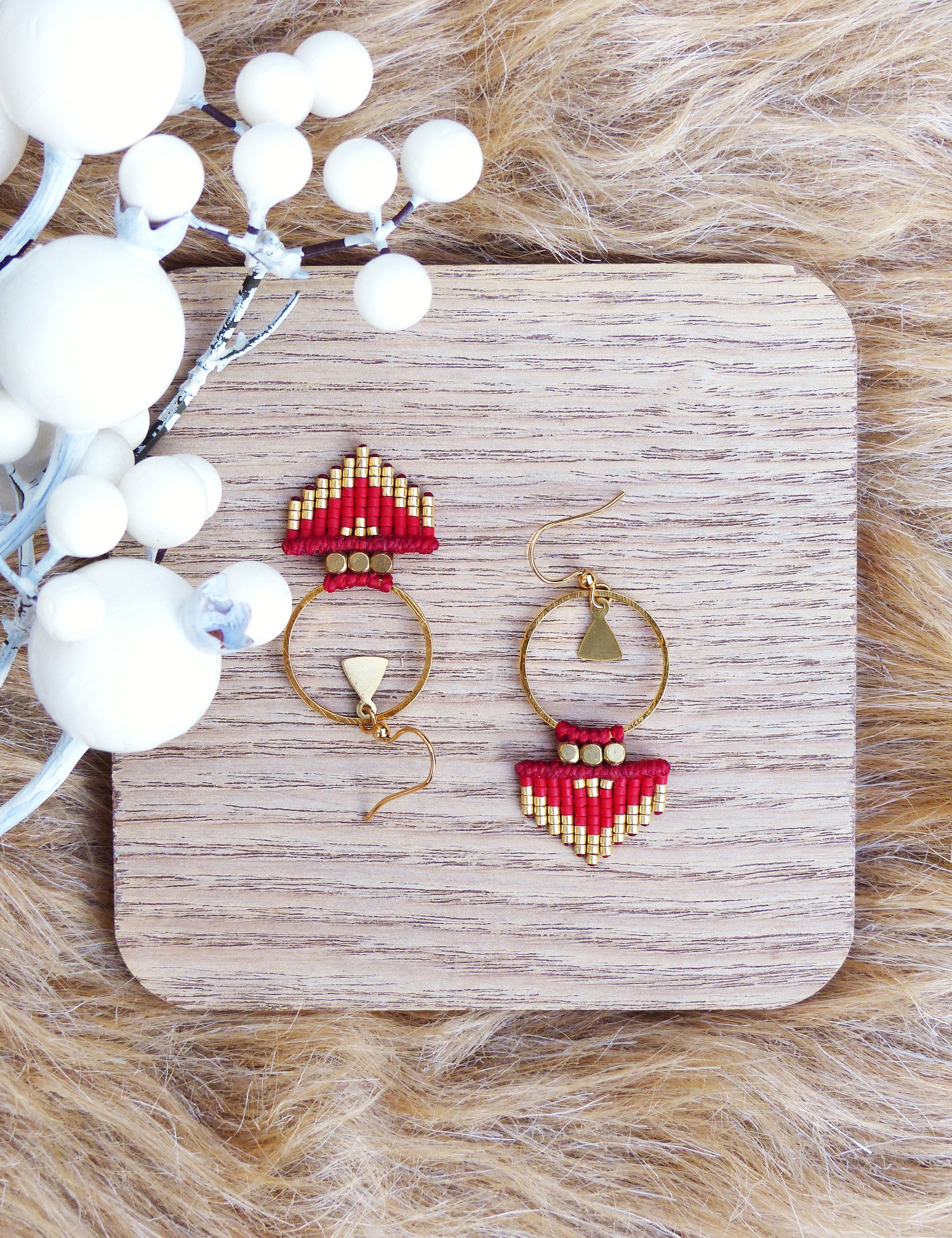 Peut inclure: Une paire de boucles d'oreilles en perles rouges et dorées avec un motif géométrique. Les boucles d'oreilles sont suspendues à une surface en bois.