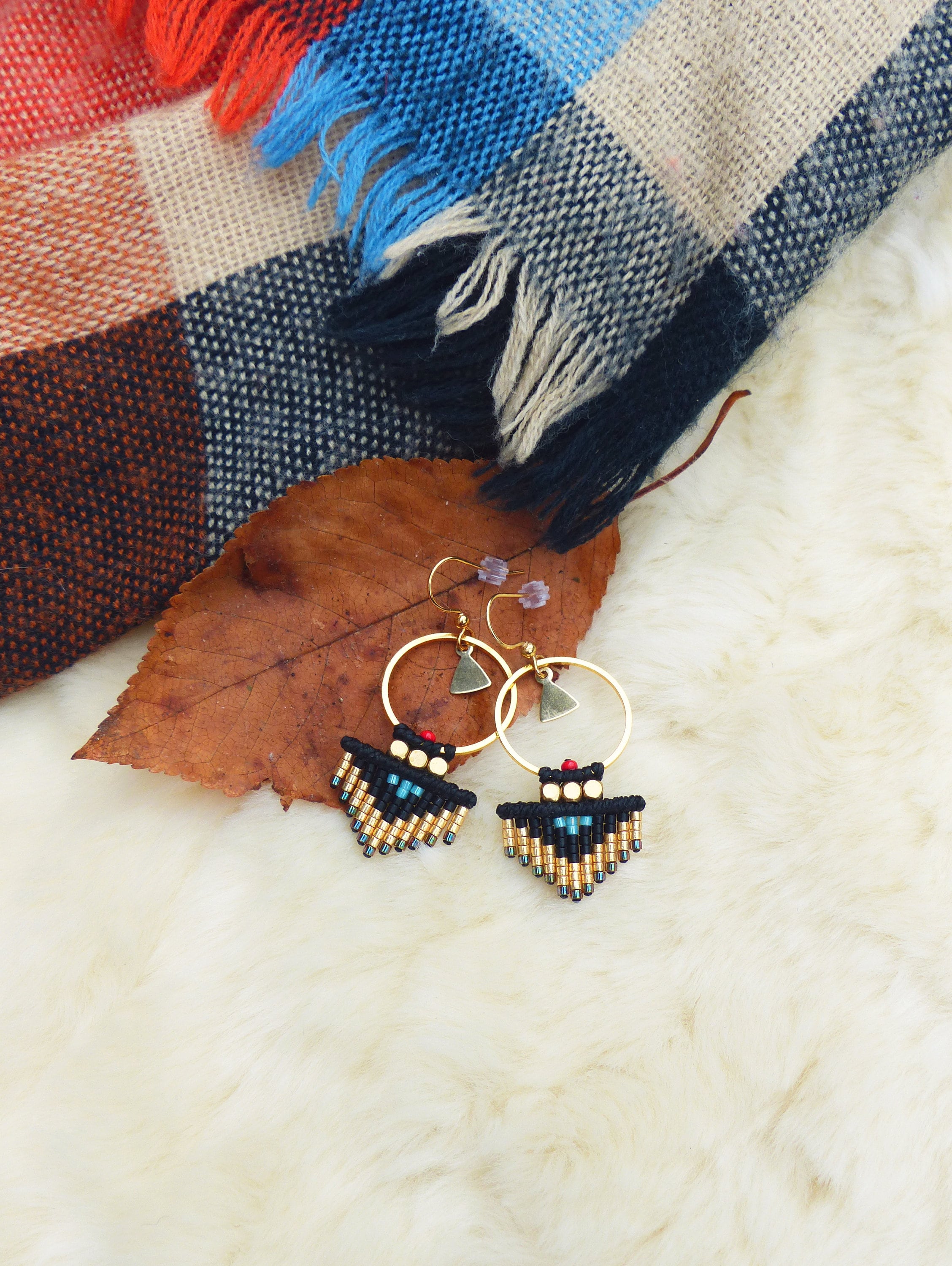 Peut inclure: Une paire de boucles d'oreilles créoles dorées avec des franges de perles noires et dorées. Les boucles d'oreilles sont sur une surface blanche et moelleuse avec une feuille brune et une couverture à carreaux en arrière-plan.