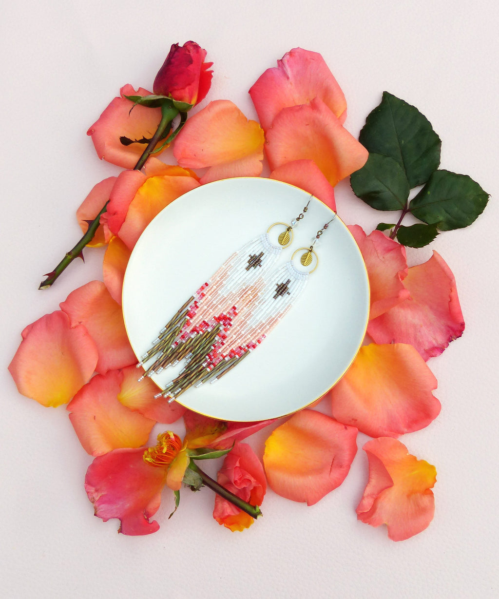 Peut inclure: Une paire de longues boucles d'oreilles en perles avec un motif rose, blanc et or. Les boucles d'oreilles sont présentées sur une assiette blanche entourée de pétales de rose.