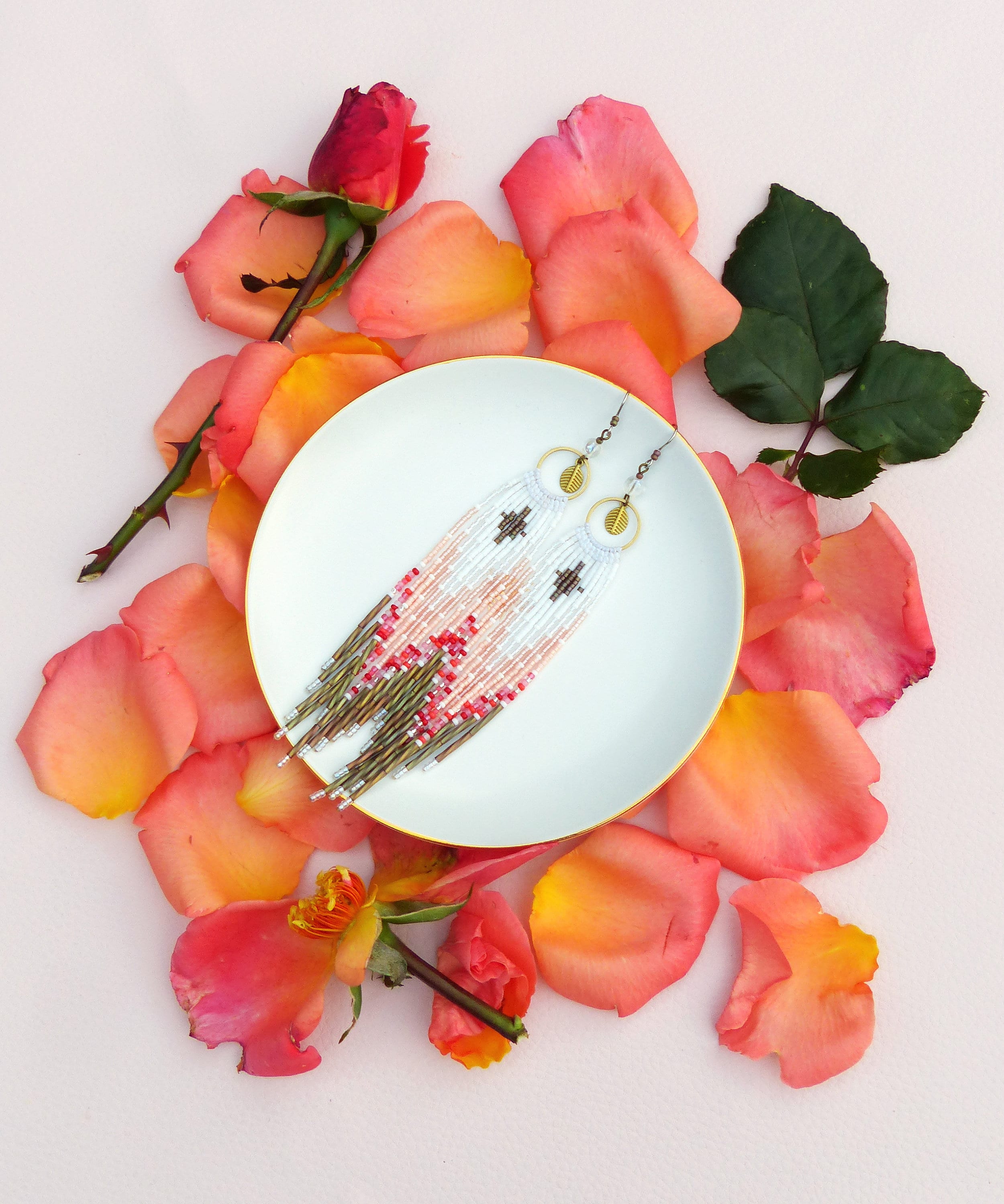 Peut inclure: Une paire de longues boucles d'oreilles en perles avec un motif rose, blanc et or. Les boucles d'oreilles sont présentées sur une assiette blanche entourée de pétales de rose.