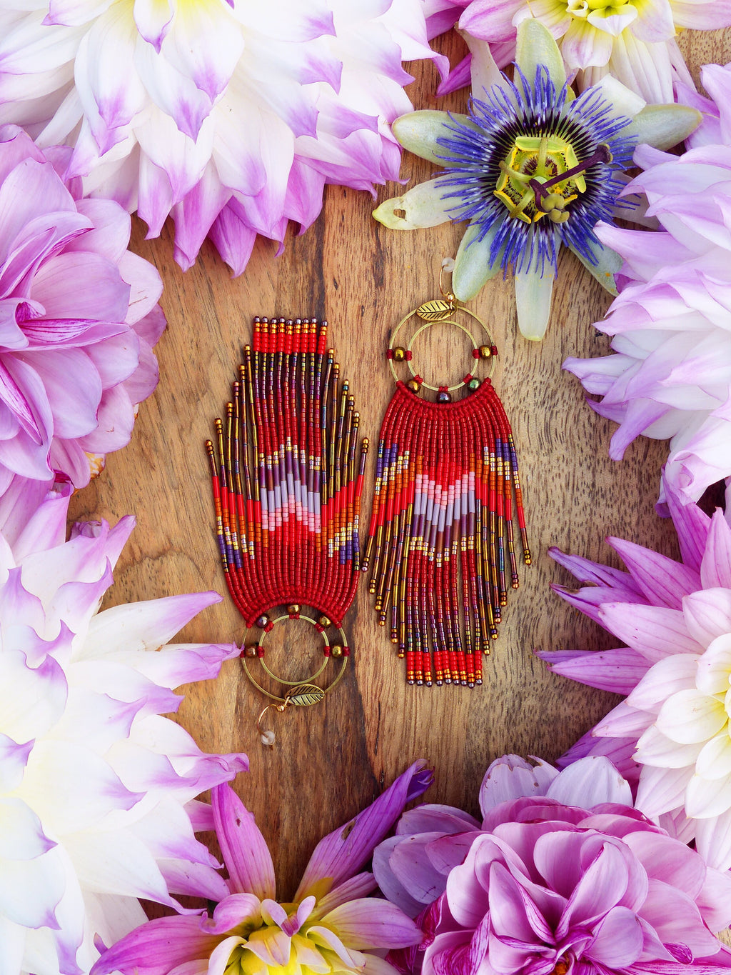 Peut inclure: Une paire de longues boucles d'oreilles rouges en perles avec des accents dorés. Les boucles d'oreilles sont suspendues à une surface en bois entourée de fleurs roses.