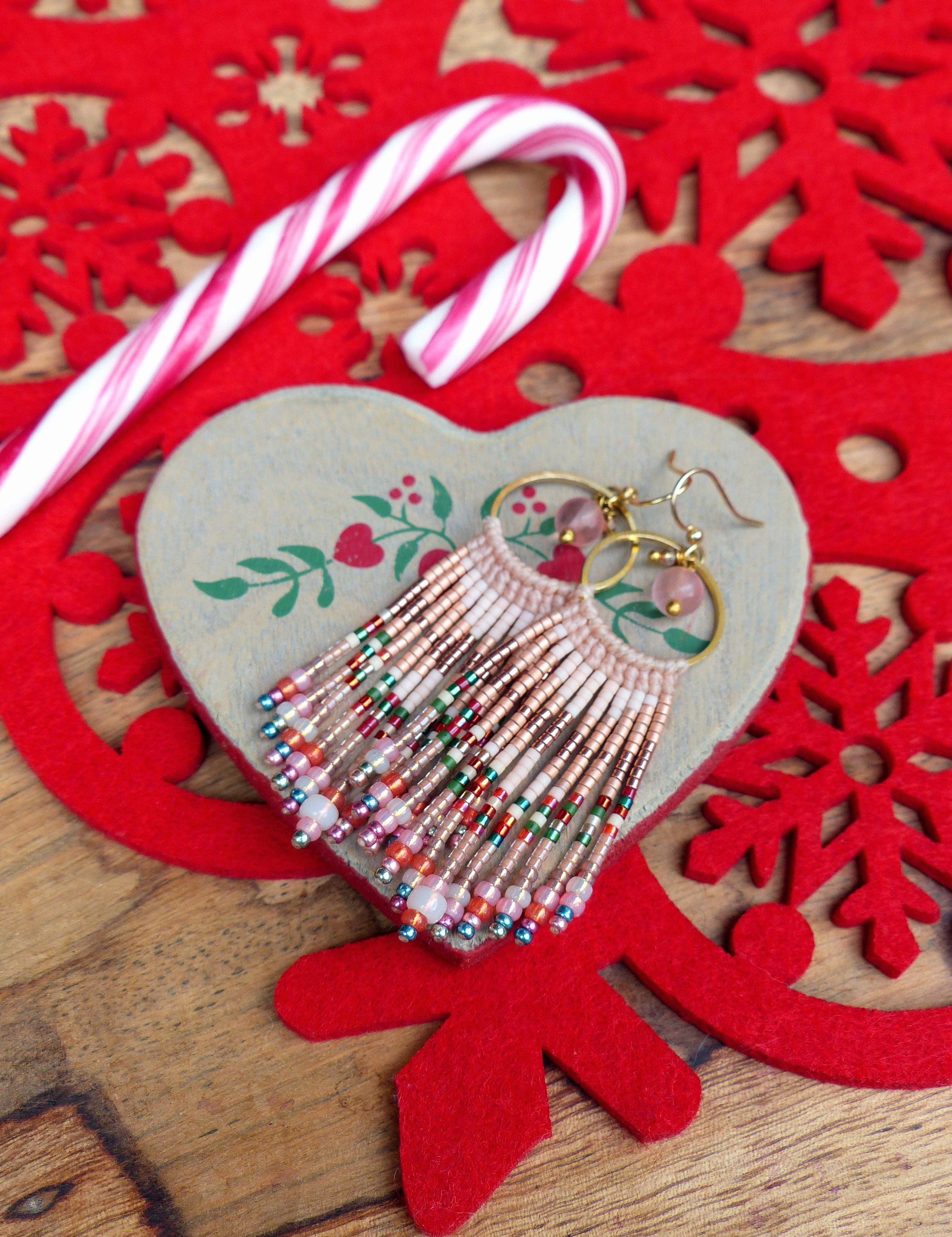 Peut inclure: Une paire de boucles d'oreilles en perles roses et brunes avec des cercles dorés et des accents de pierres roses. Les boucles d'oreilles sont présentées sur une surface en bois en forme de cœur avec un fond de flocon de neige rouge.