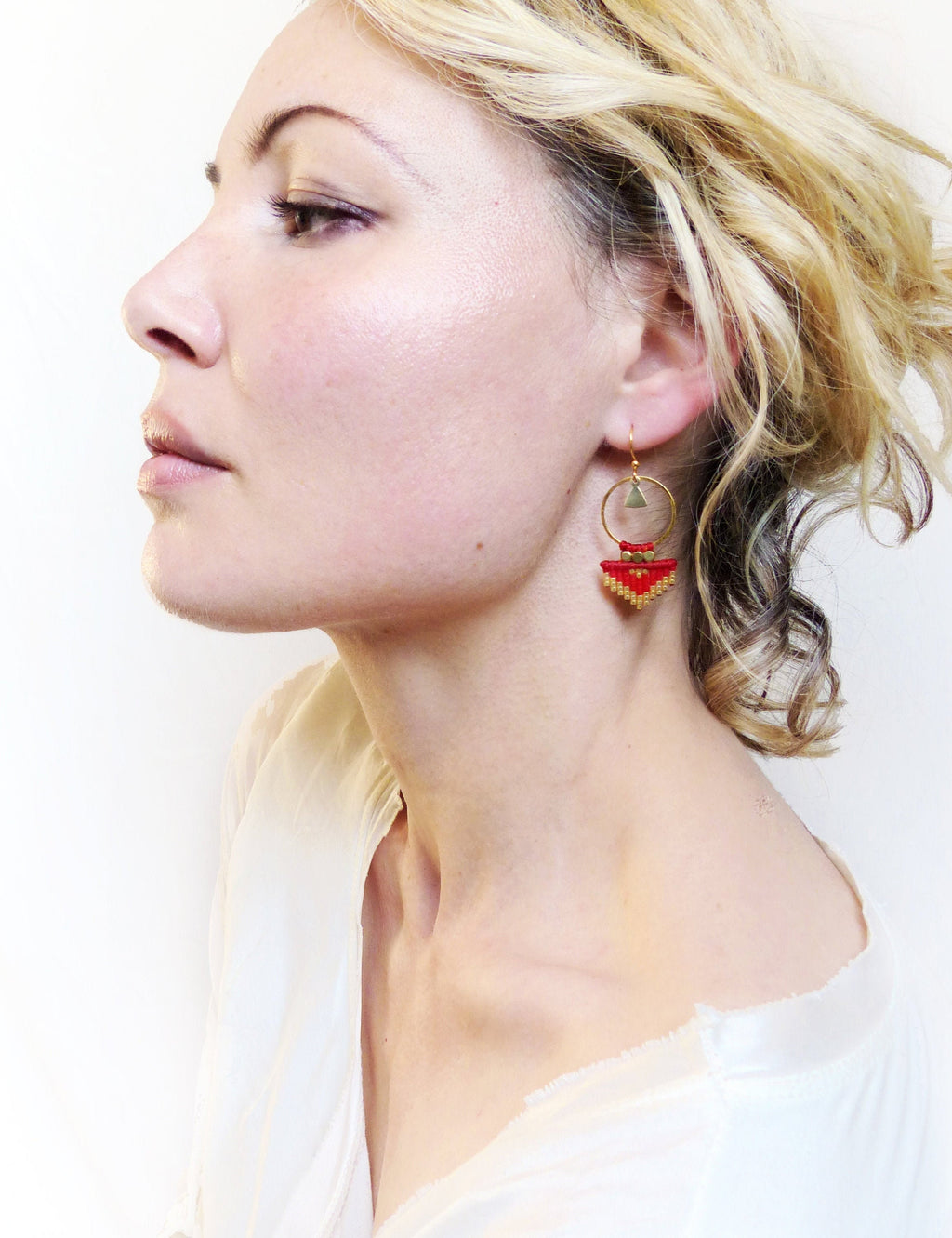 Peut inclure: Une paire de boucles d'oreilles créoles dorées avec un pendentif géométrique rouge. Le pendentif est fait de petites perles rouges et a une forme triangulaire.